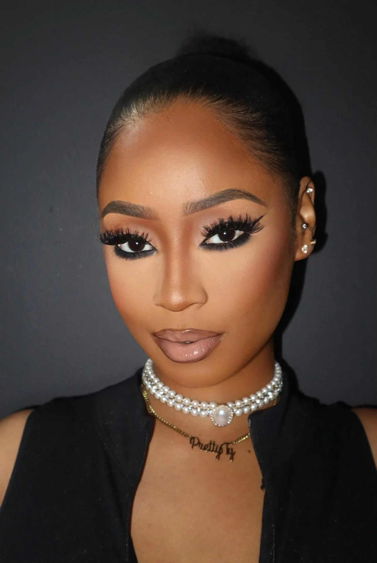 Close-up portrait of a woman with styled hair, prominent makeup, pearl necklace, and earrings, against a dark background.