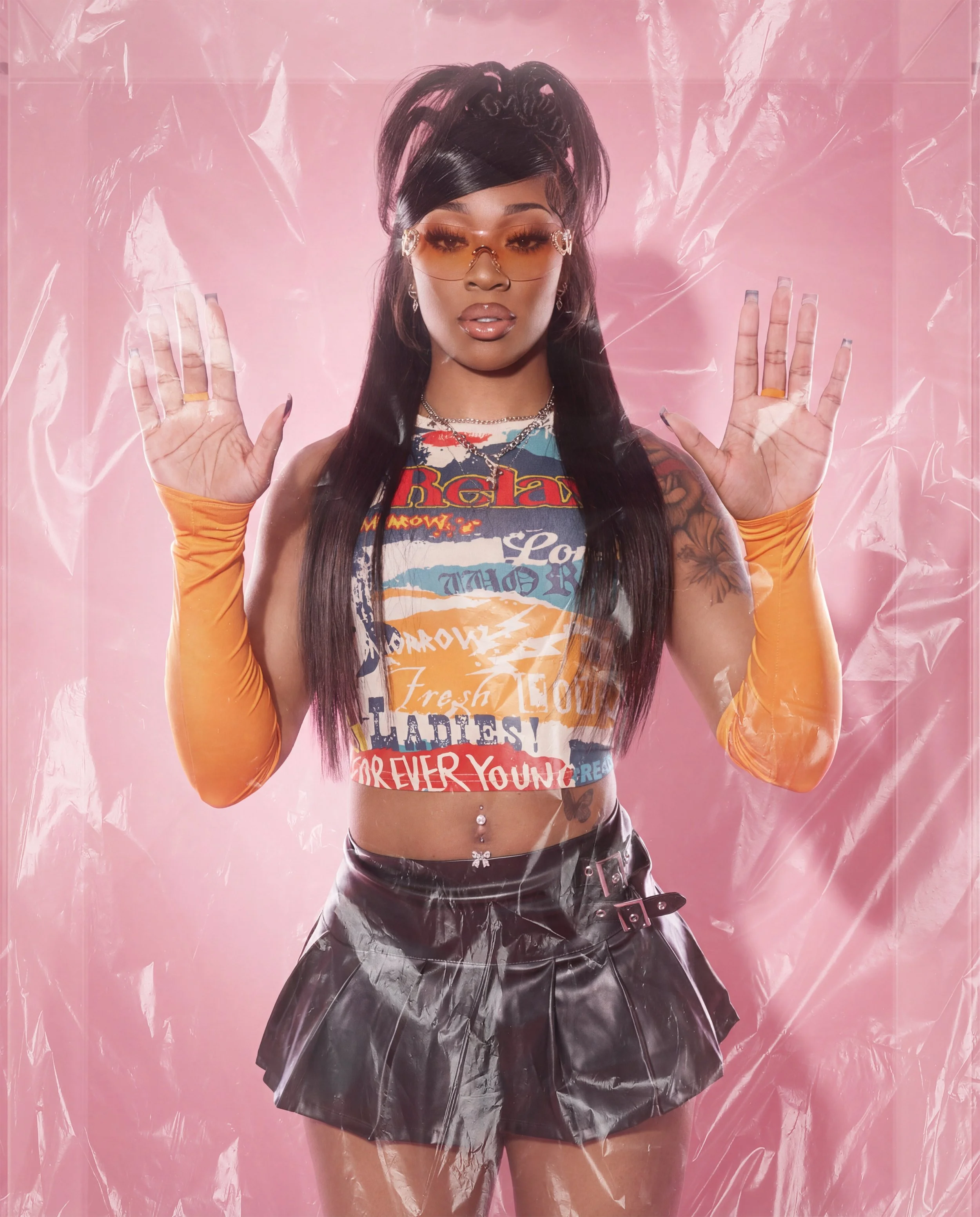 A woman with long dark hair, wearing orange gloves, sunglasses, a patterned crop top, and a black leather skirt, standing behind a pink background covered with plastic and pressing her palms against it.