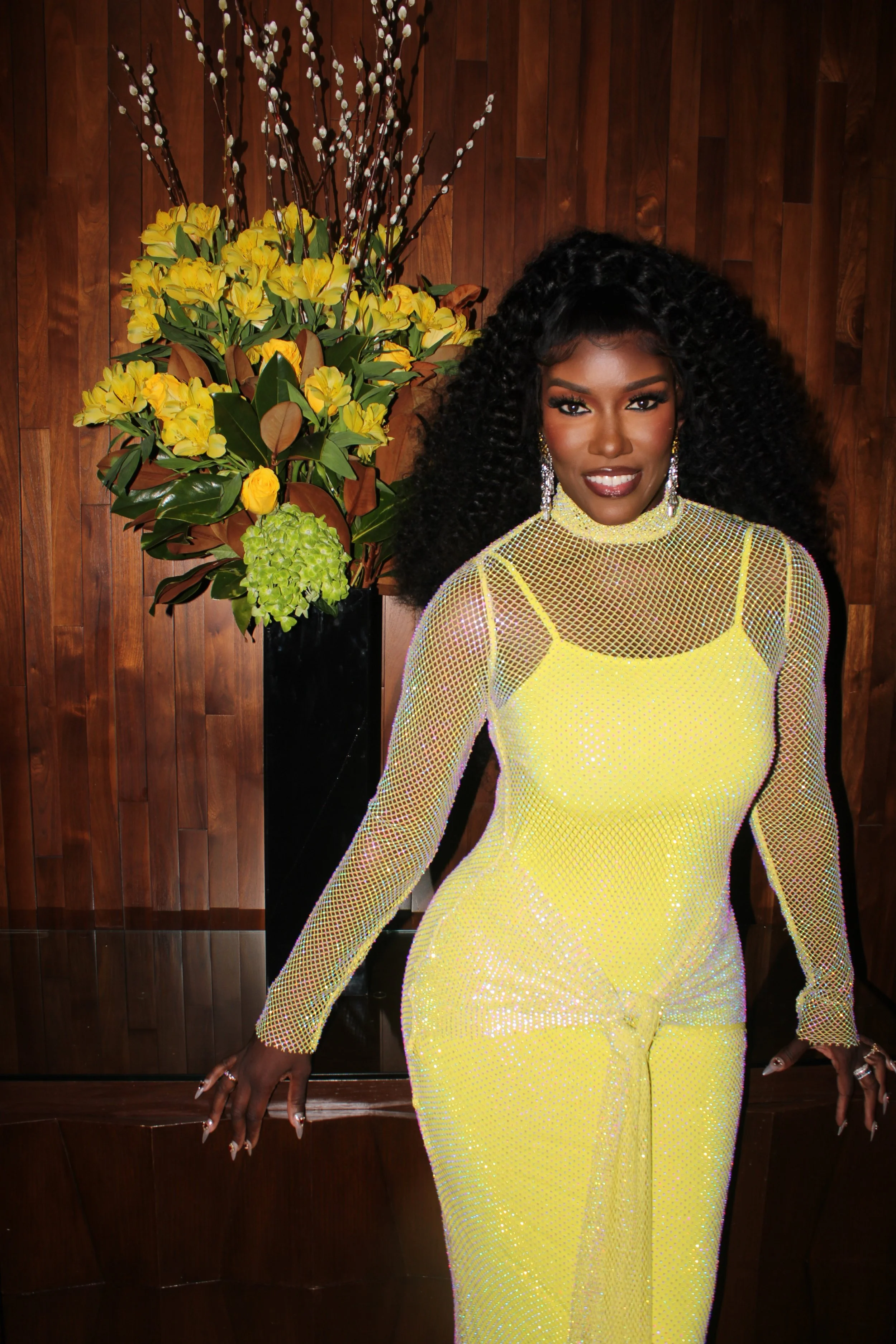 A woman with dark skin and long curly black hair wearing a sparkly yellow dress with sheer mesh sleeves, standing in front of a wooden wall and a large flower arrangement with yellow and green flowers.