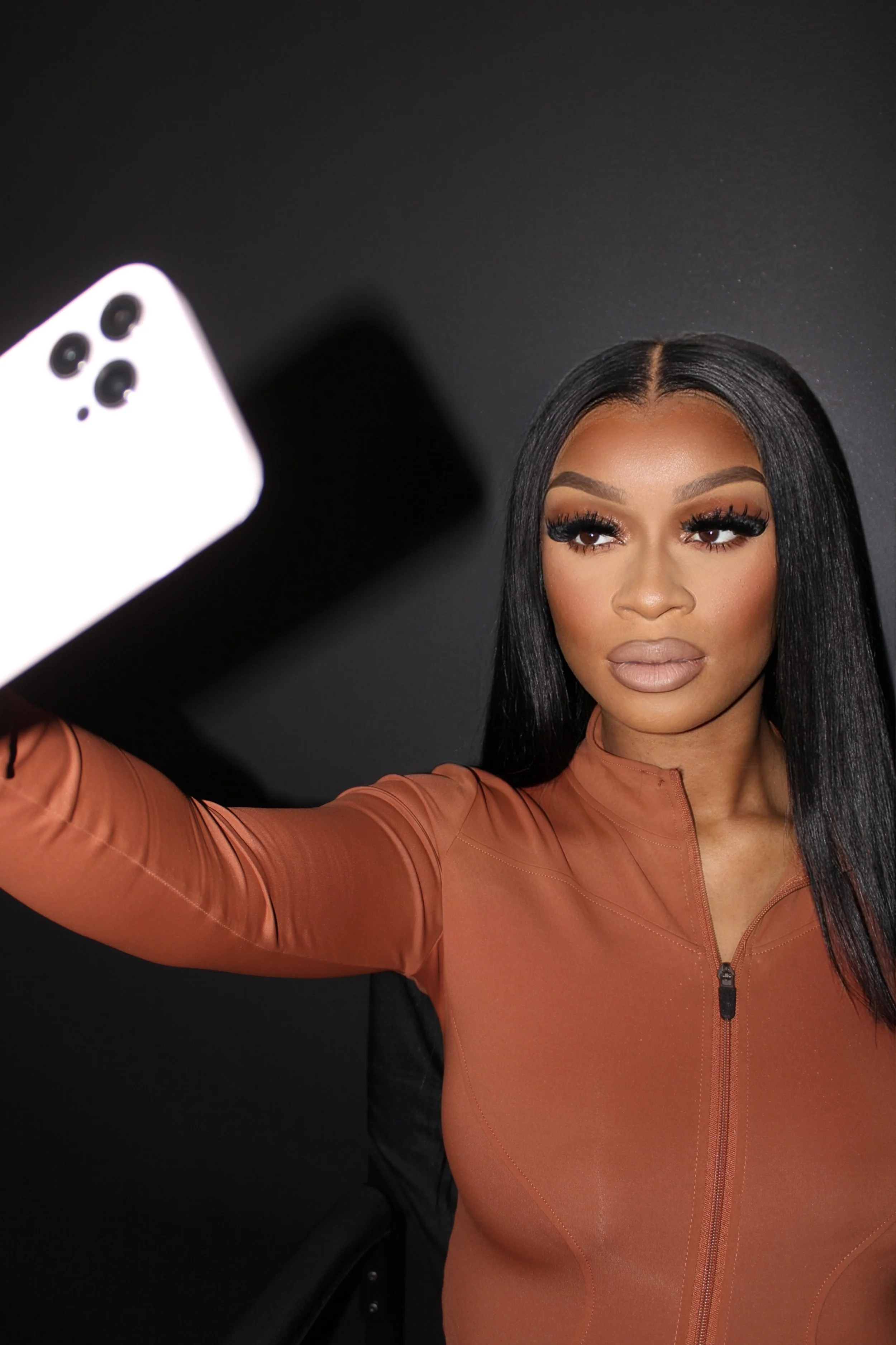 A woman with long black hair, makeup, and wearing a fitted orange zip-up jacket is taking a selfie with a white smartphone against a dark background.