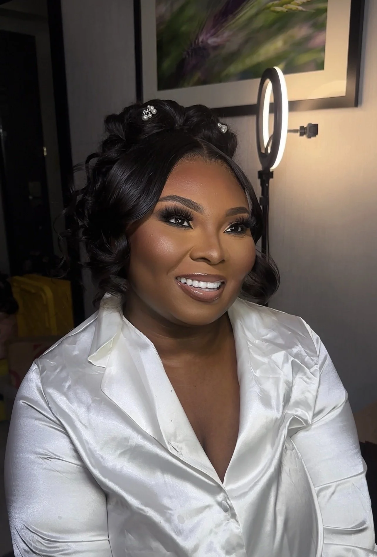 A woman with styled hair and makeup, smiling, seated in front of a ring light and a wall with a framed picture.