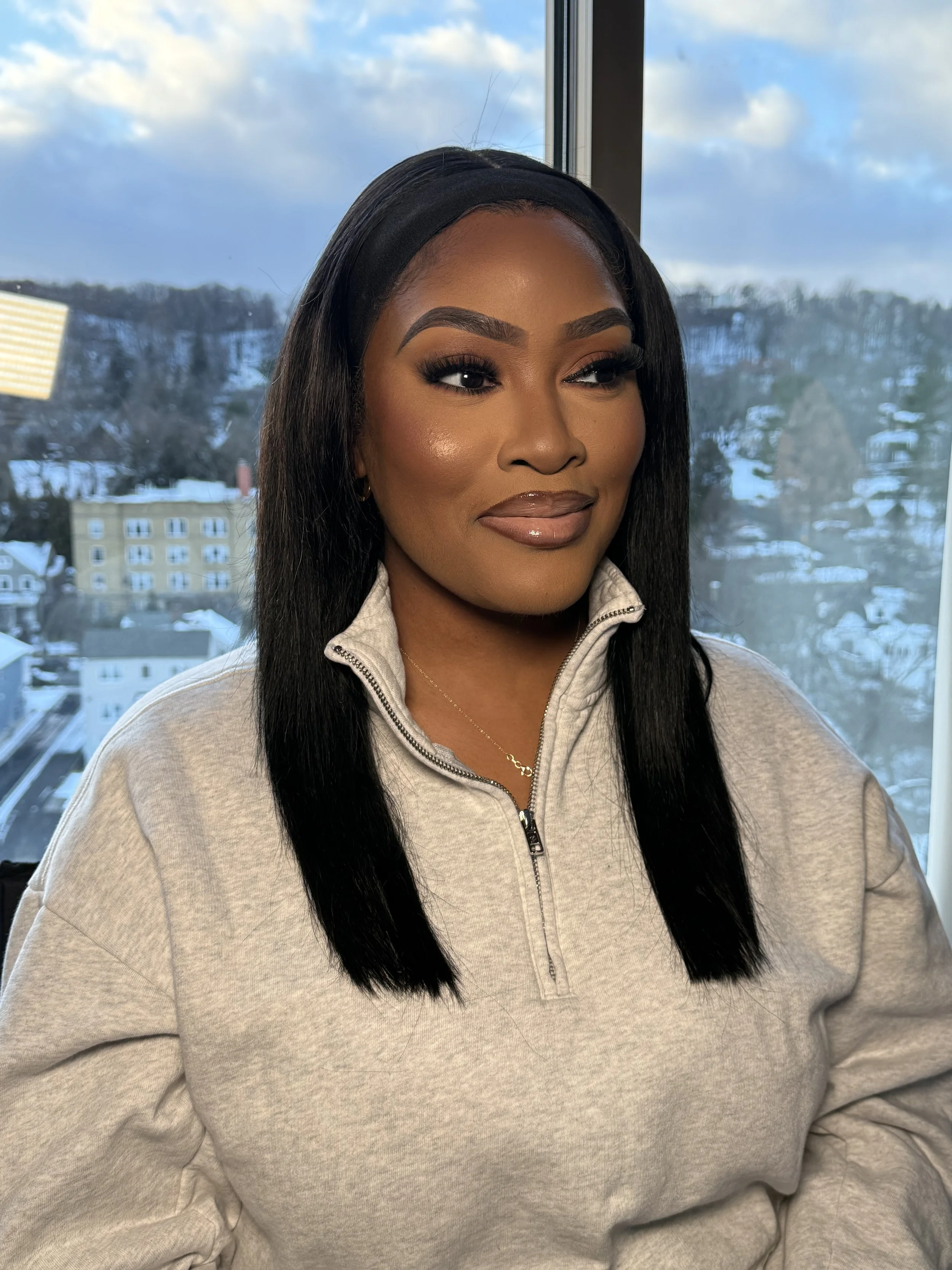 A woman with long black hair, wearing makeup, a gray zip-up sweatshirt, and a gold necklace, smiling softly with a winter cityscape background seen through a window.