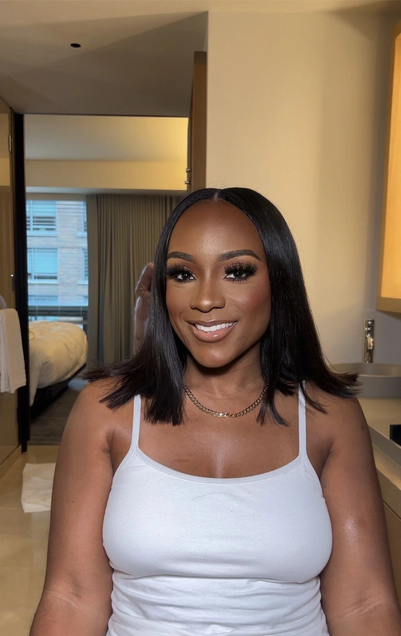 A woman with shoulder-length black hair, wearing a white tank top and a gold chain necklace, smiling inside a hotel room with a bed and balcony in the background.