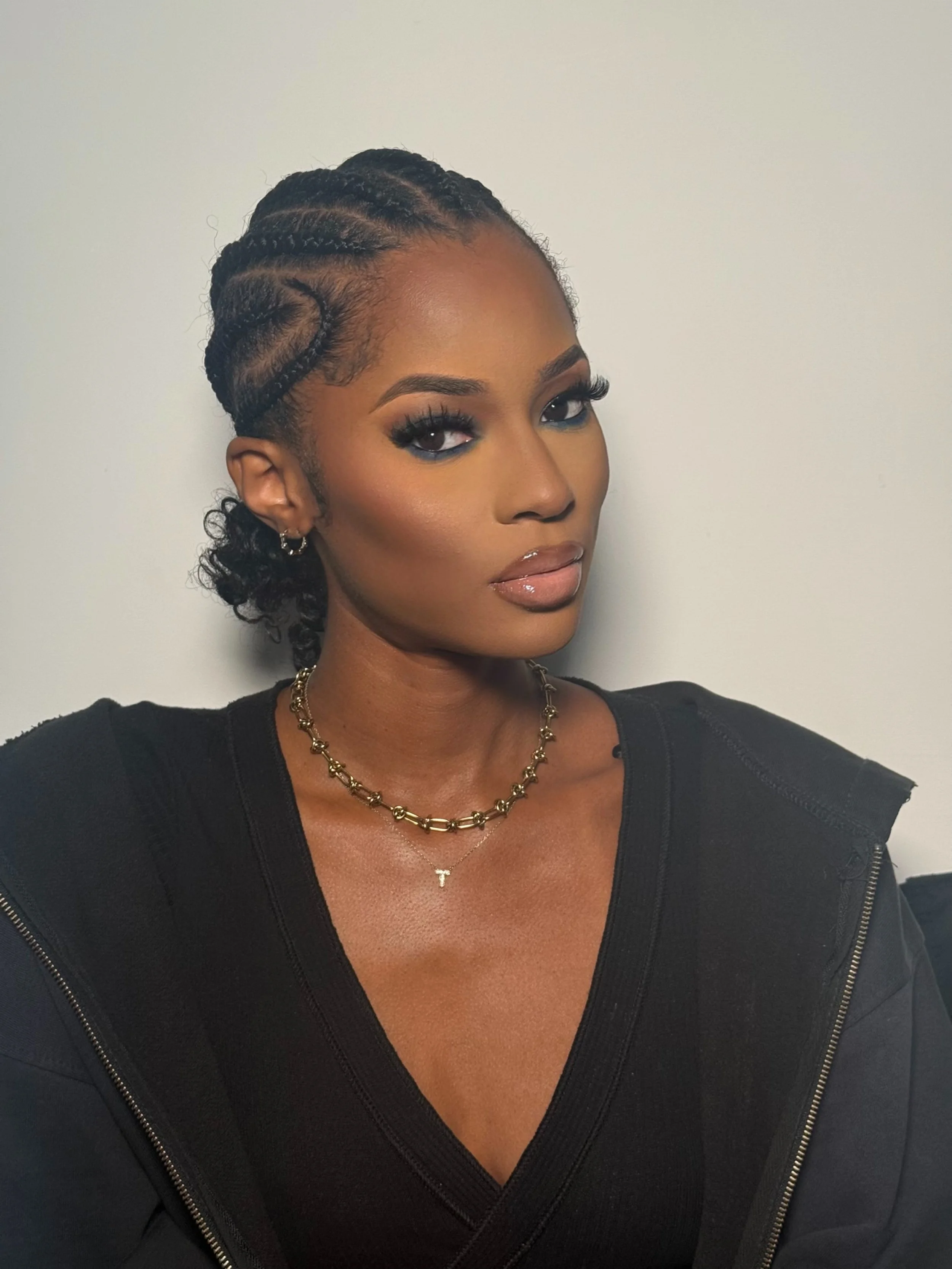 Portrait of a woman with braided hair, tattoos on her chest, wearing a black top and gold jewelry, looking at the camera.