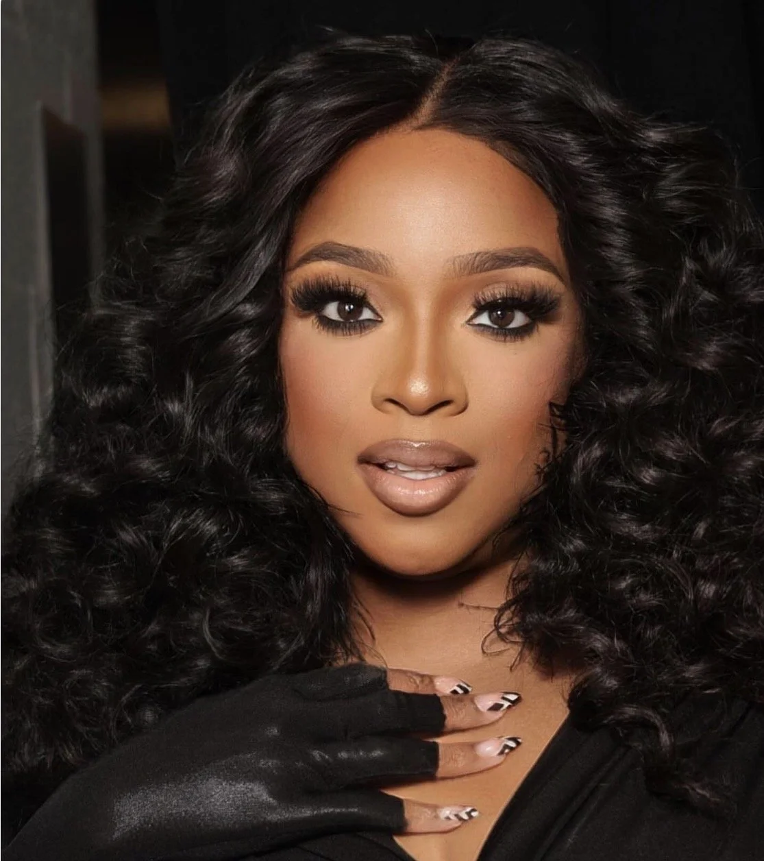 Close-up of woman with voluminous curly black hair, makeup, and nail art, looking at the camera.