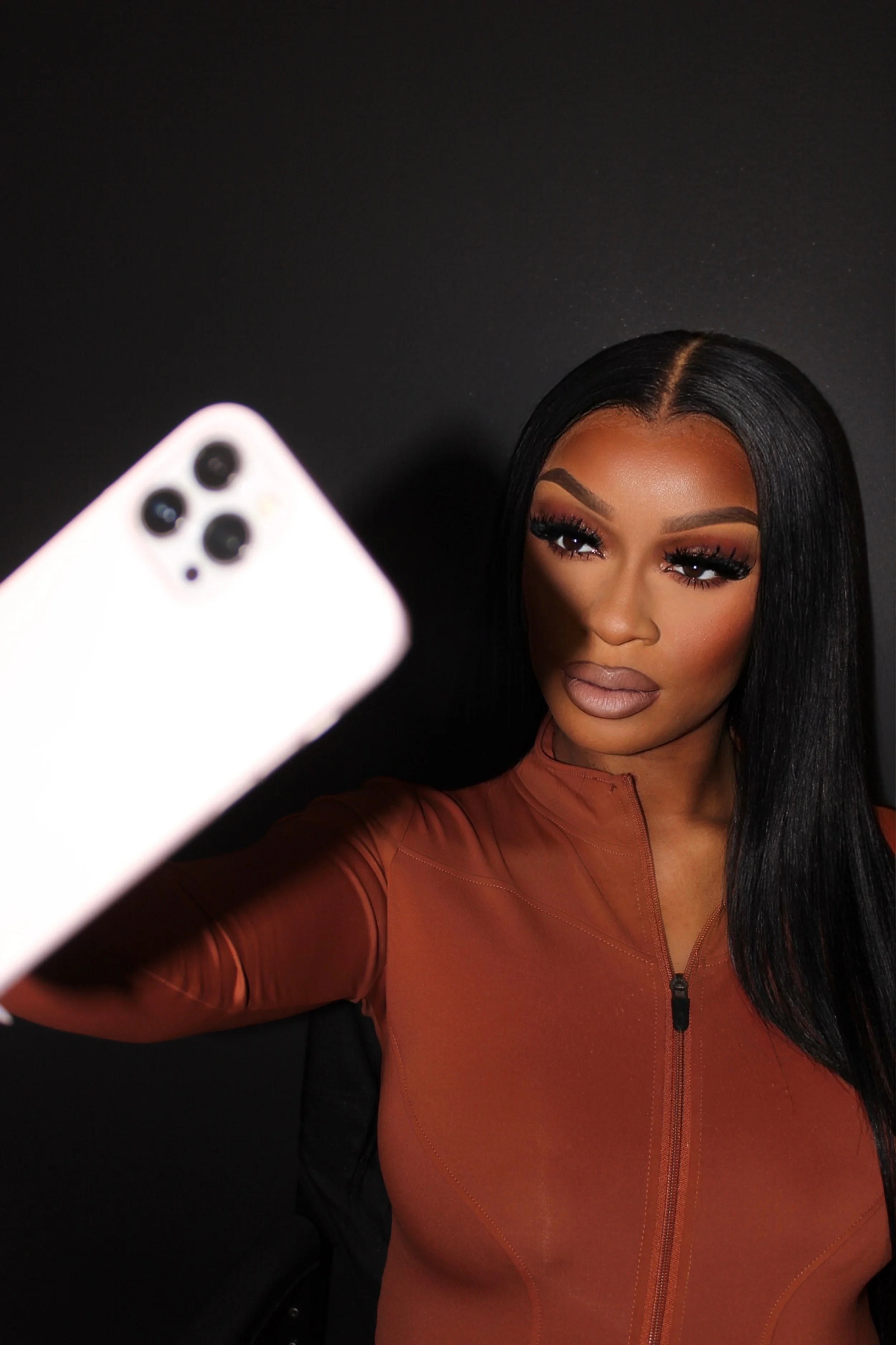 A woman with long black hair taking a selfie with a smartphone, against a black background.