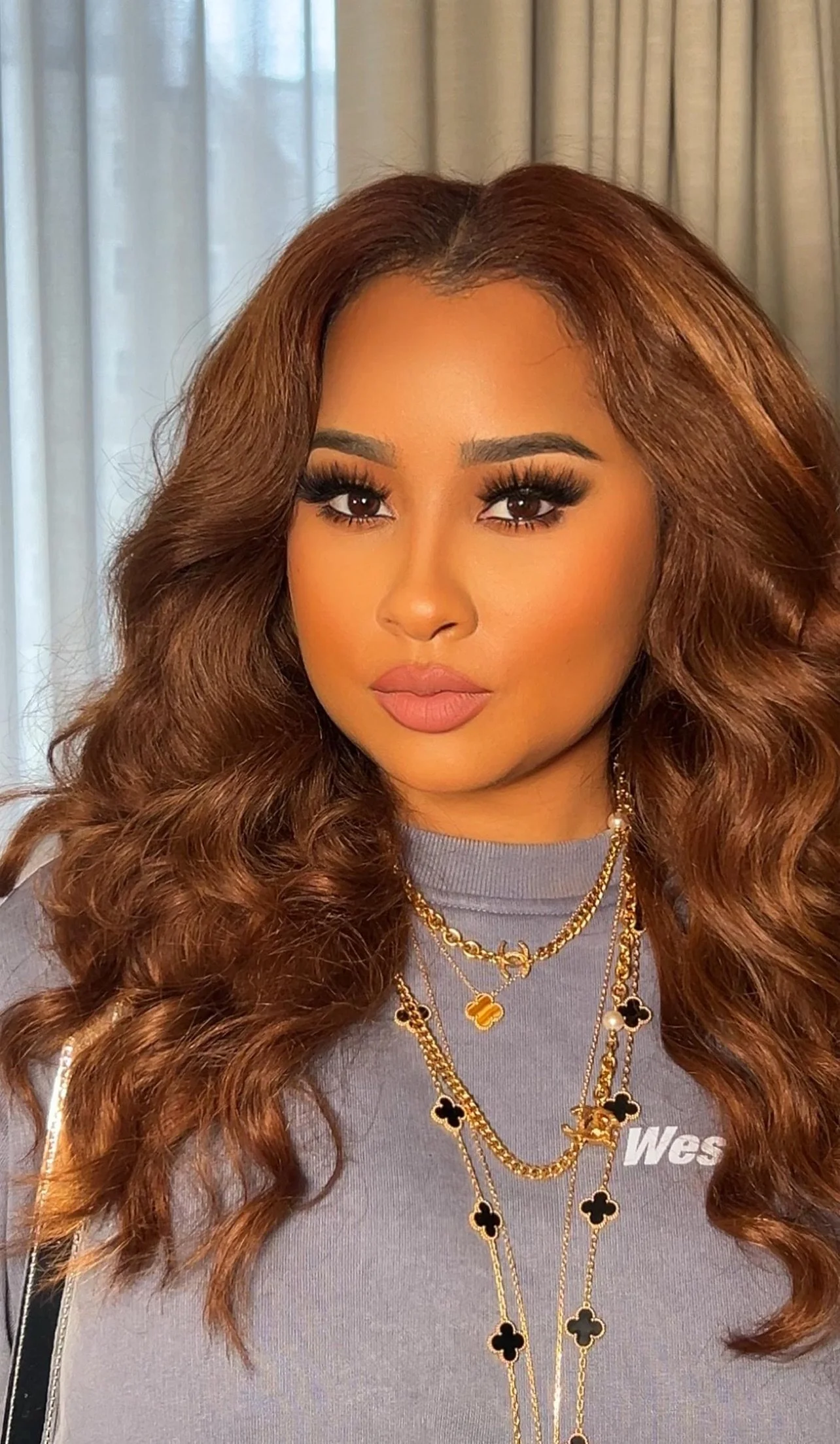 A woman with long, curly, reddish-brown hair, wearing multiple gold necklaces with black and gold flower pendants, a gray shirt with 'West' printed on it, posing indoors with curtains in the background.