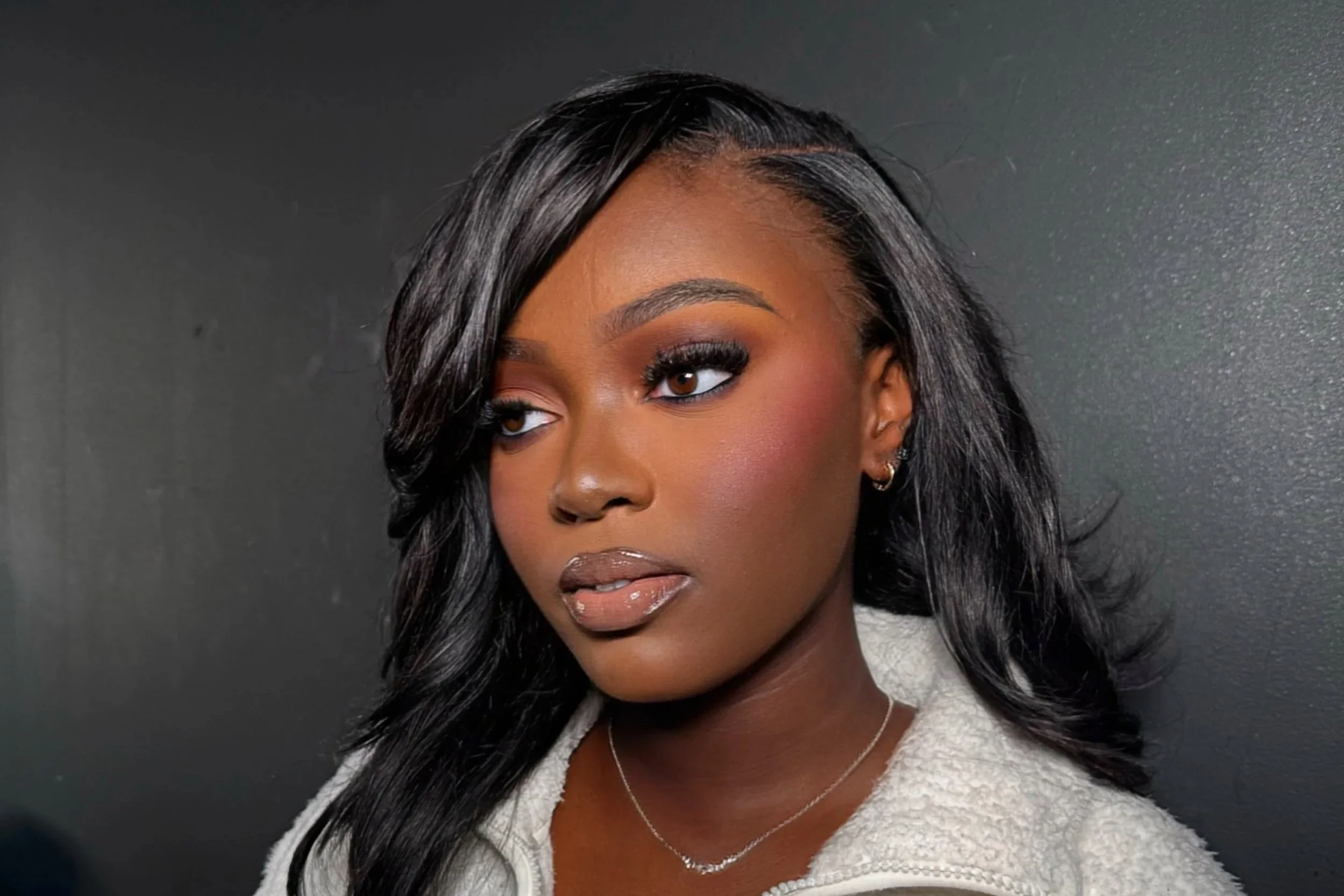 A young woman with dark hair styled in loose waves, wearing makeup with dark eyeshadow, false eyelashes, and glossy lips, dressed in a light-colored textured top, standing against a dark background.