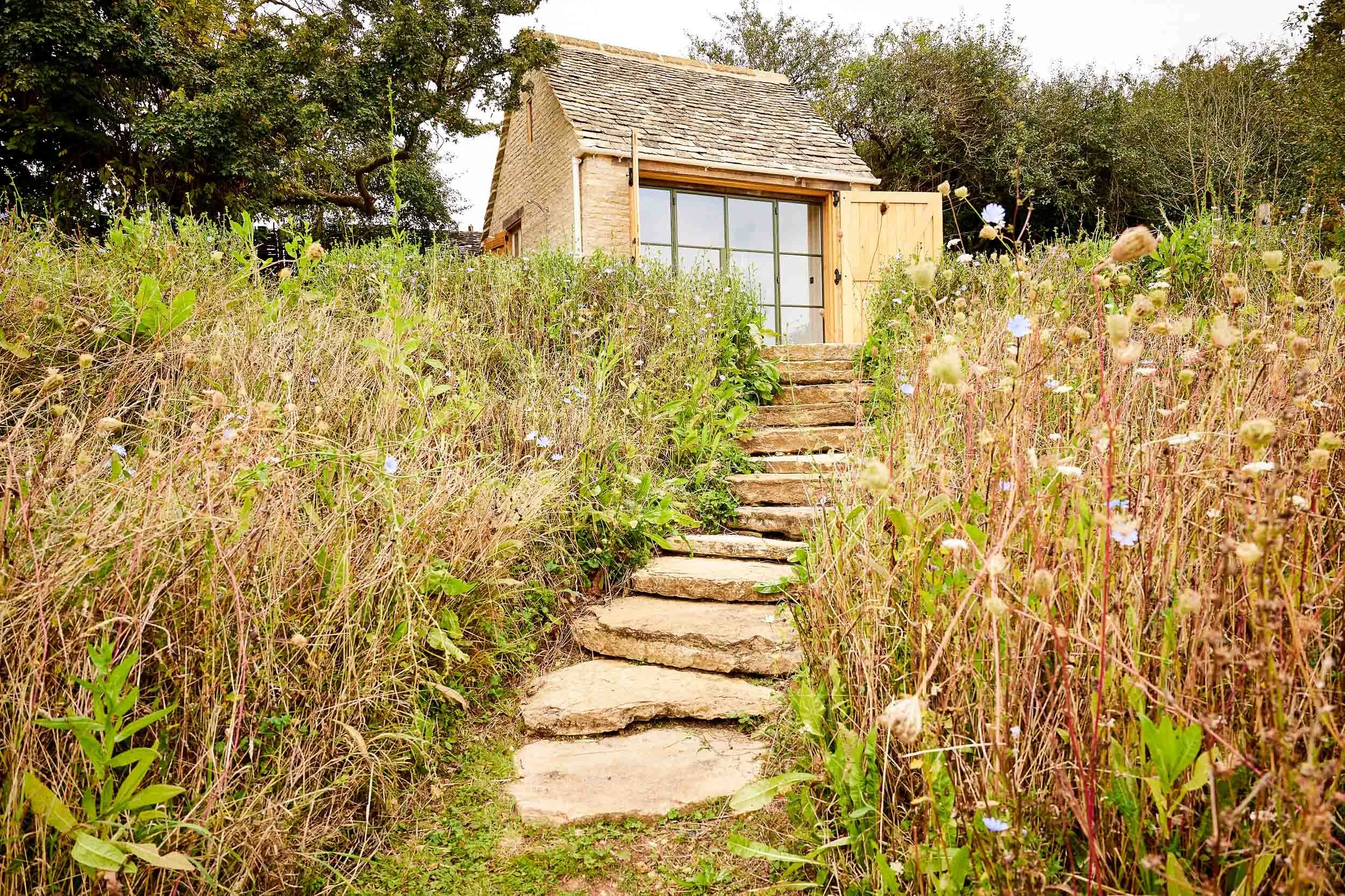 A rustic stone pathway leading up a hill through tall wildflowers to a small cottage with large glass windows and a wooden door, surrounded by trees.