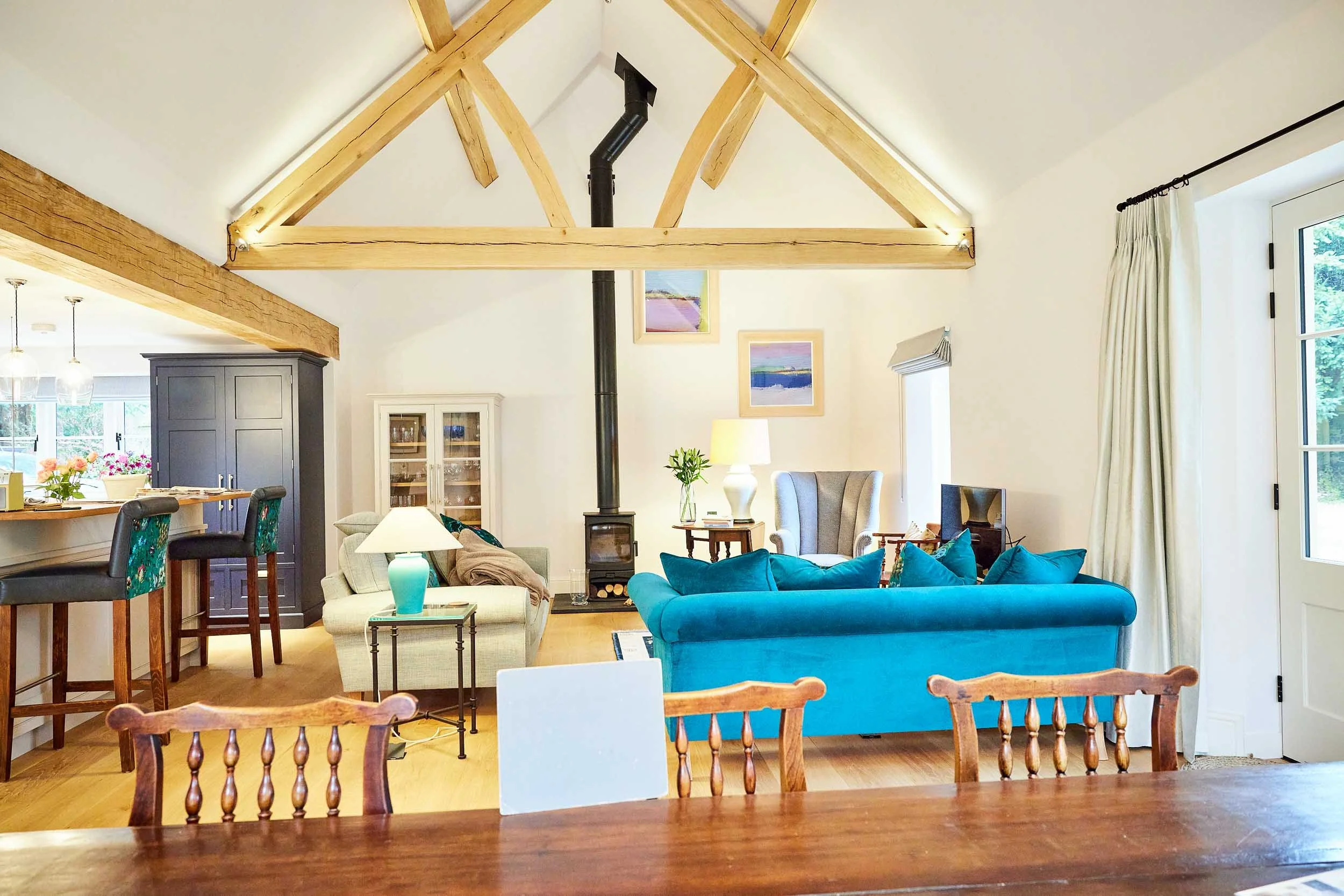 Bright living room with vaulted ceiling, exposed wooden beams, a black stove pipe, and colorful artwork on the wall. Features include a teal sofa, a beige armchair, side tables with lamps, and a TV. Large windows with curtains let in natural light.
