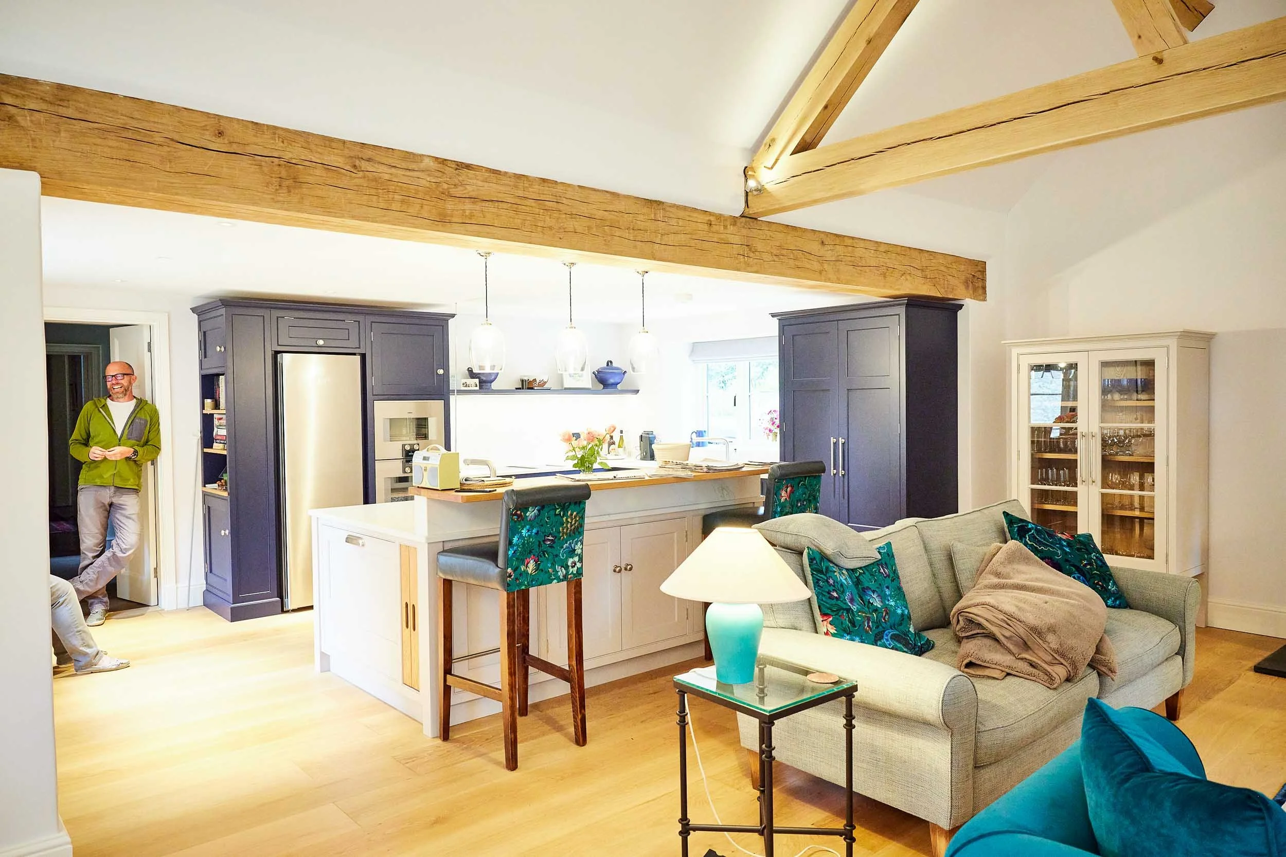 Interior of a living room and kitchen with wooden beams, navy blue cabinetry, a beige sofa with patterned pillows, a glass side table with a turquoise lamp, and two men standing near the kitchen.