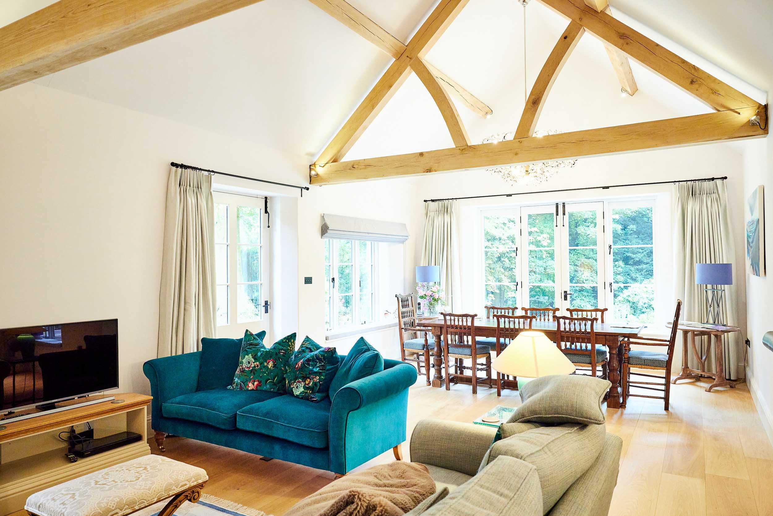 Bright living room with vaulted ceiling, exposed wood beams, large windows, a blue sofa with floral pillows, beige armchair, wooden dining table with chairs, television, and side tables with lamps.
