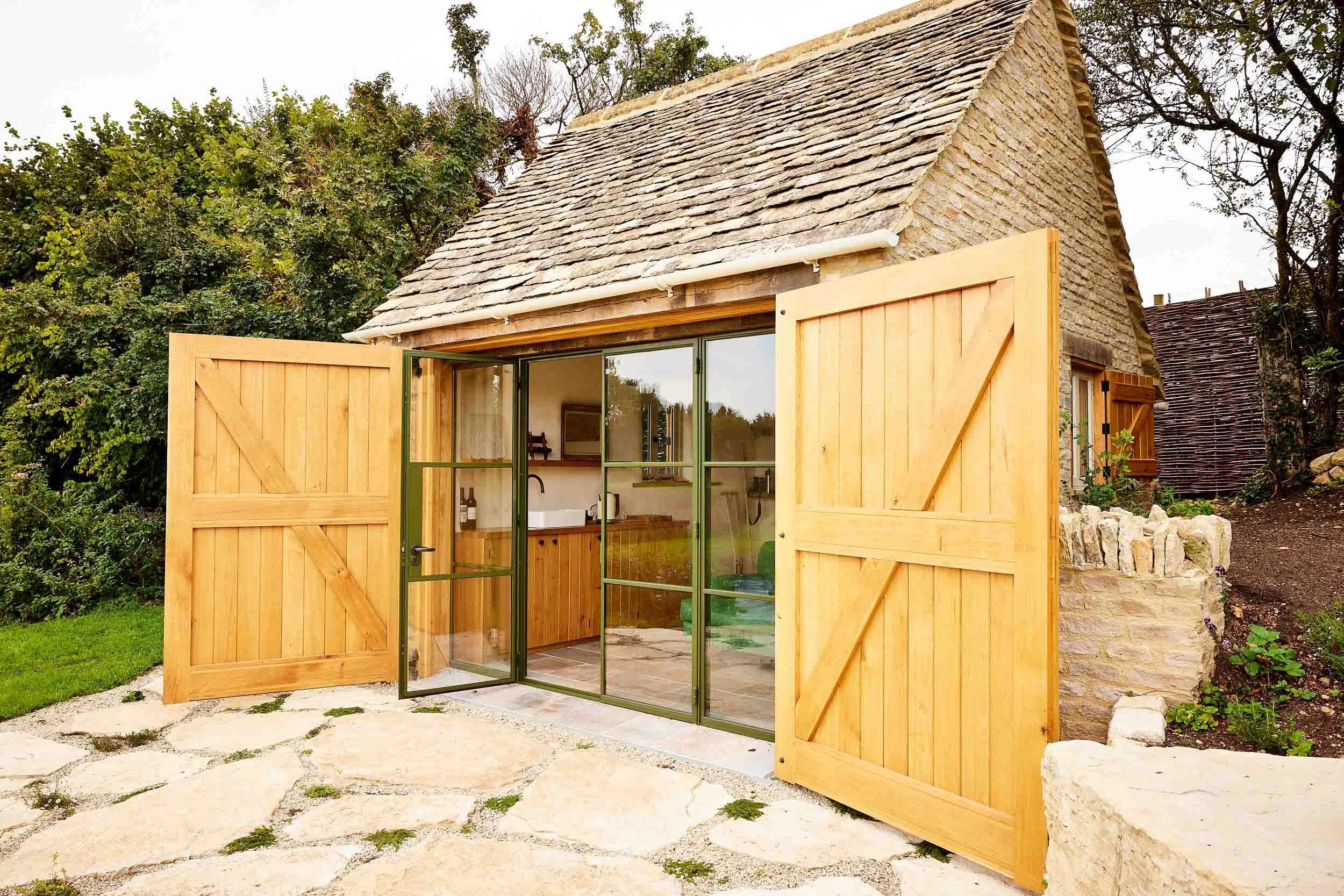 Small stone cottage with open wooden barn-style doors and a sliding glass door revealing a rustic kitchen interior.