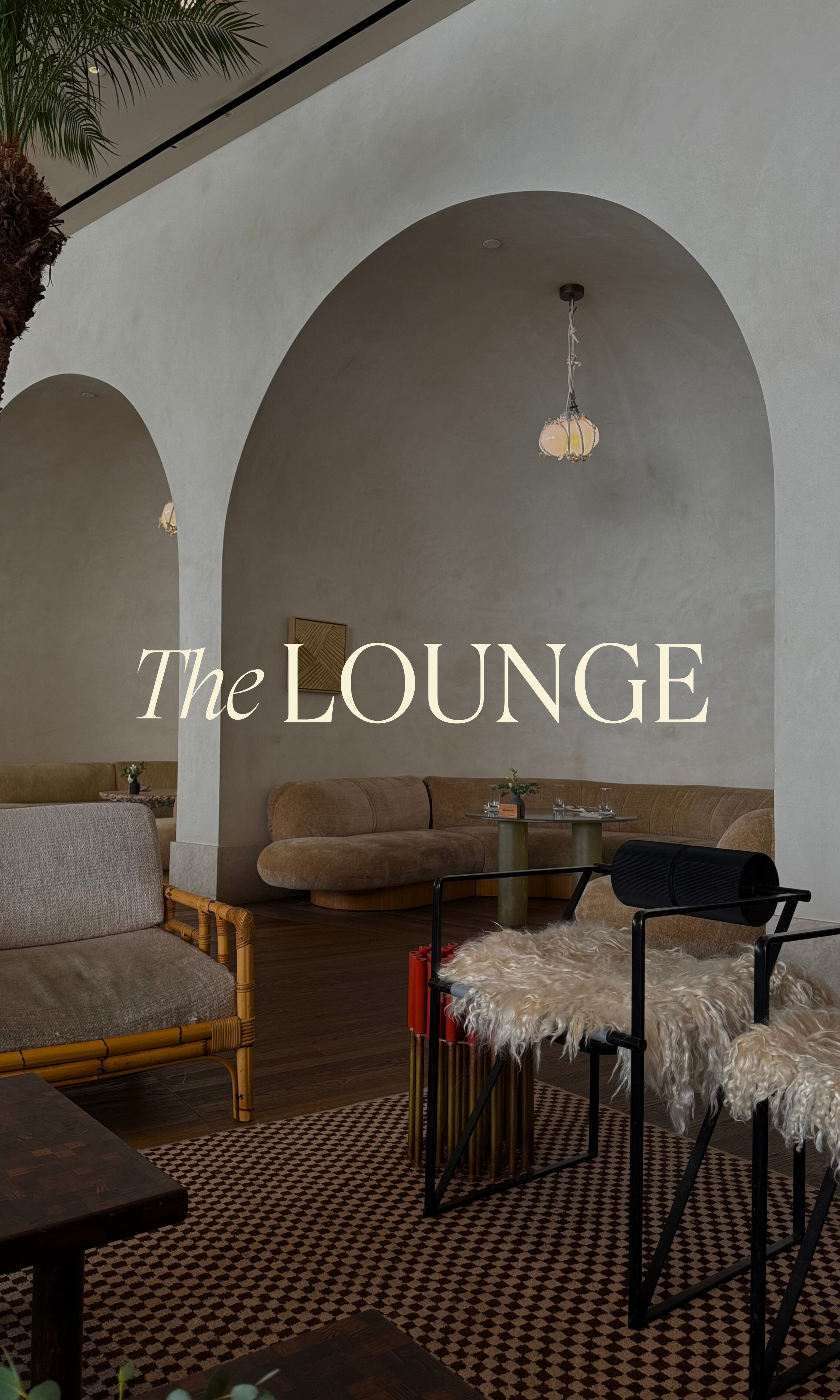 Interior of a cozy lounge with arched ceilings, beige and brown furnishings, a small table with plants, hanging light fixtures, and a patterned rug. With words "The Lounge" overlayed on top.