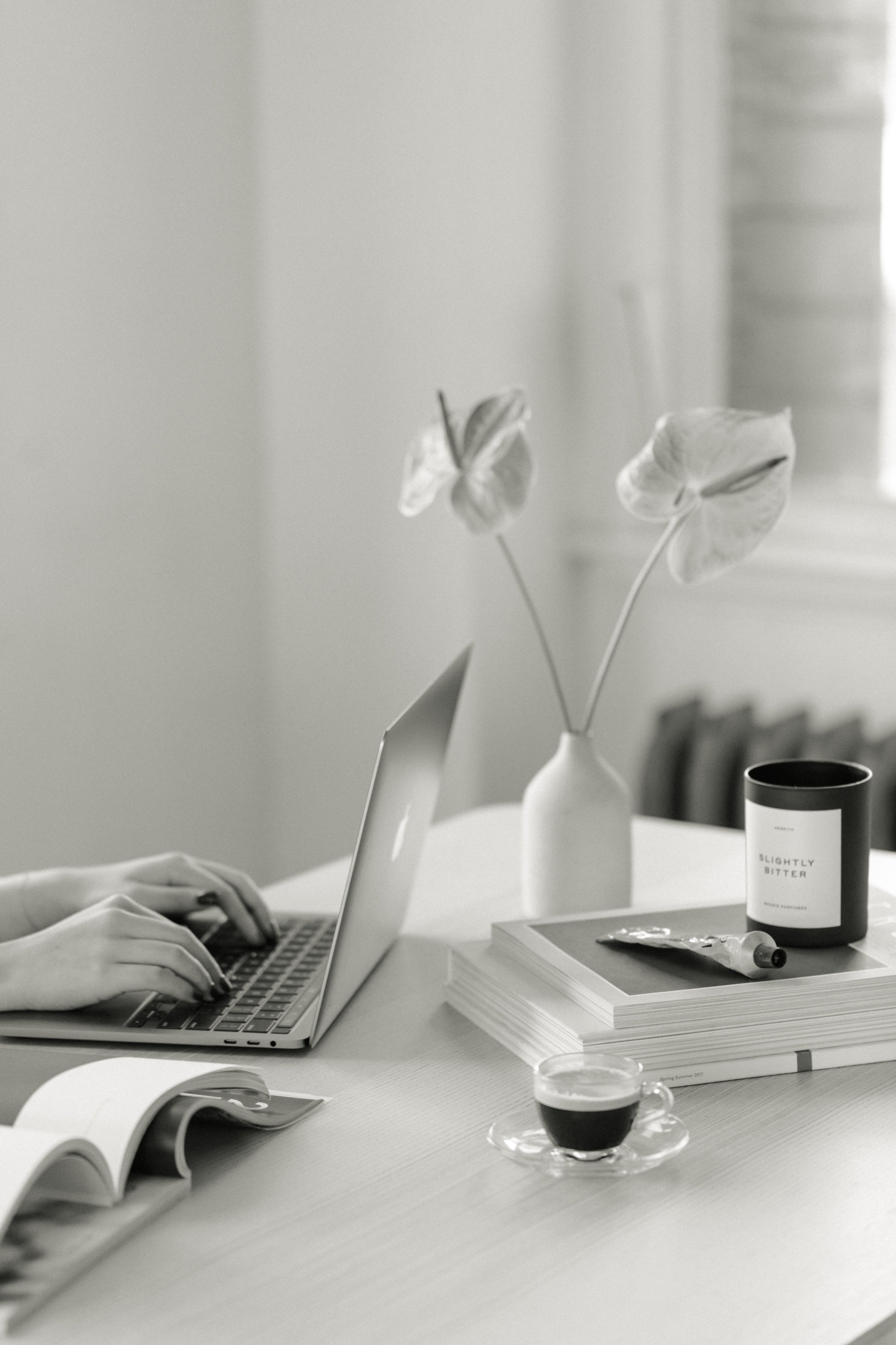 A person's hands typing on a laptop keyboard on a desk with a magazine, a coffee cup, a book, a tube of cream, a black candle labeled 'Slightly Bitter,' and a vase with two many-petaled flowers in the background.