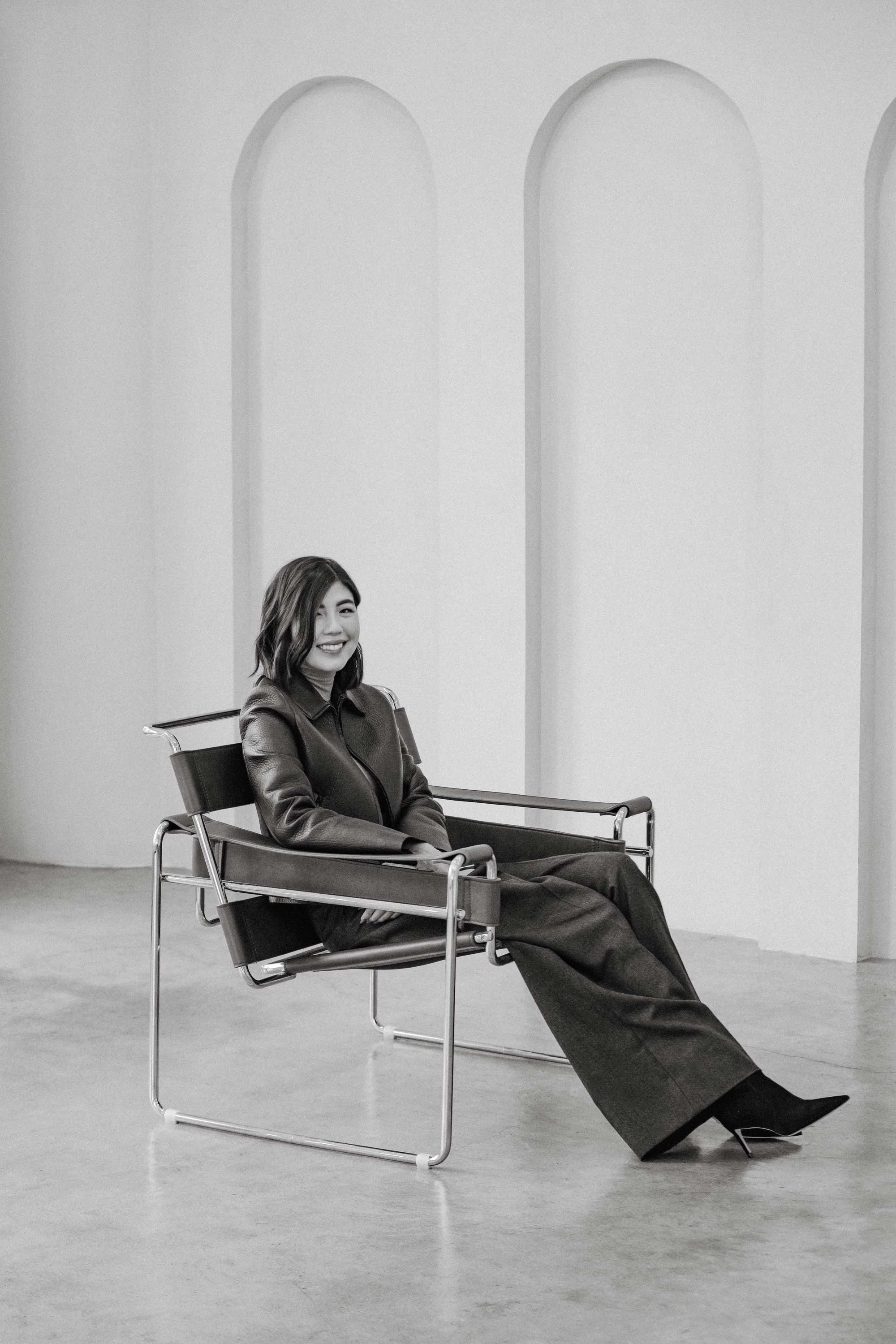 A woman sitting in a modern chair, smiling, in a minimalist room with arched doorways or windows.