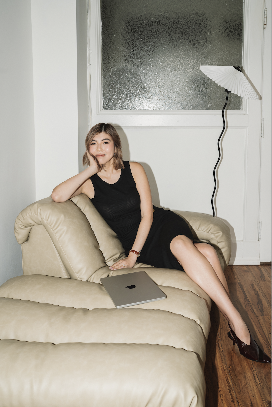 A woman in a black sleeveless dress sitting on a beige sofa, smiling with her hand resting on her cheek, next to a closed laptop, in a room with wooden floor and textured frosted glass window.