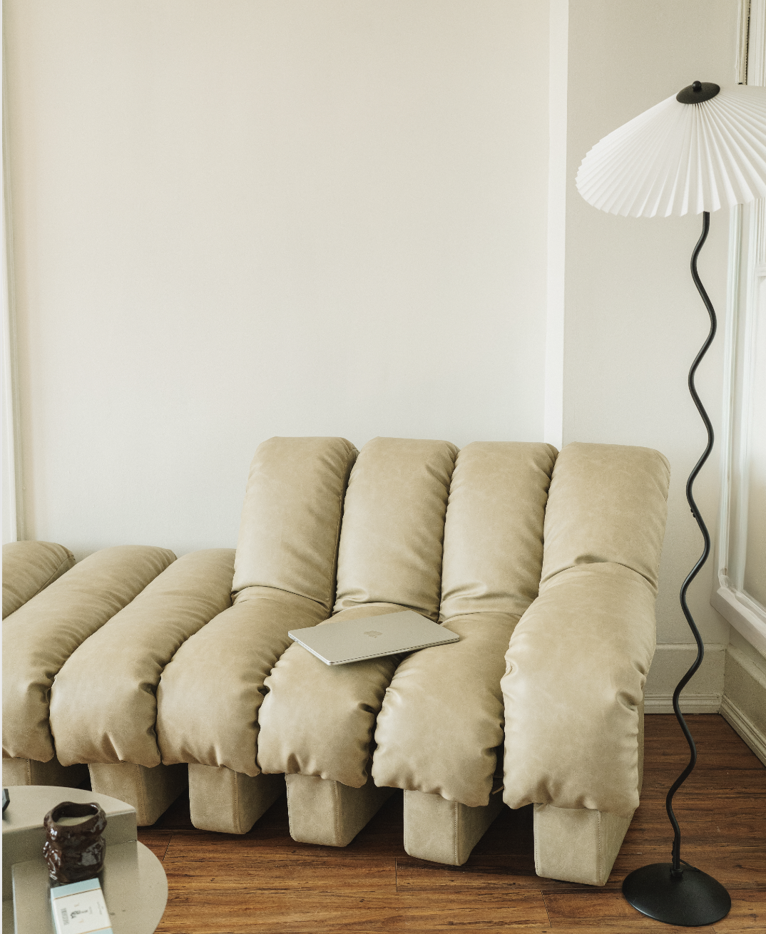 A beige sectional sofa with a closed silver laptop on it, next to a black standing lamp with a white, pleated shade, in a room with hardwood floors and a blank white wall.