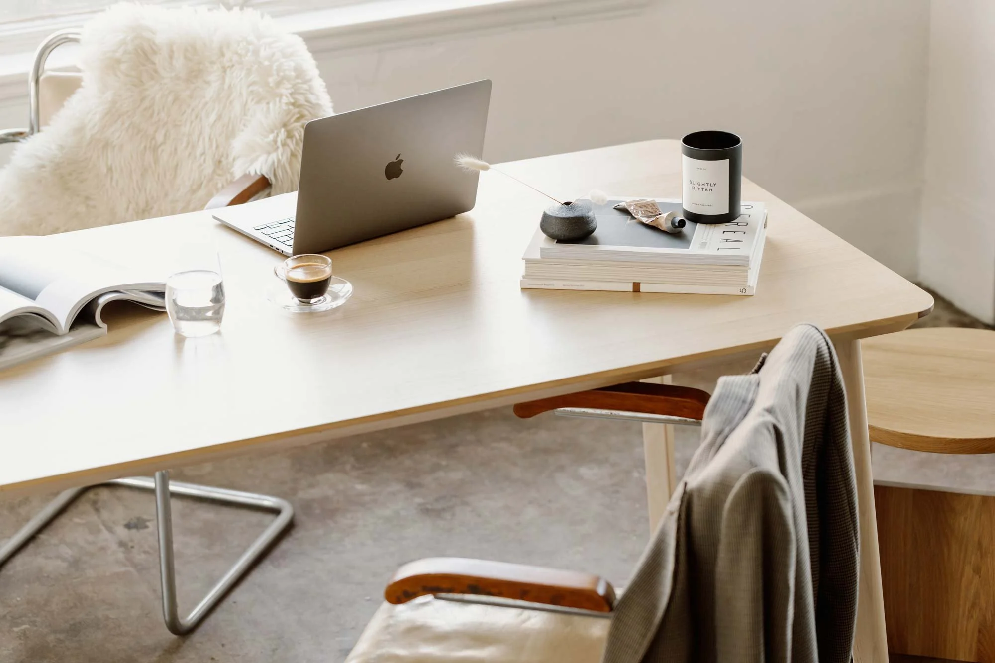 A light wooden desk with a silver Apple MacBook, an open magazine, a glass of water, a small espresso shot, and a black candle, with a stack of magazines, a stone vase, and miscellaneous items on top. A beige chair with a plaid blazer draped over it and a white fluffy blanket on the chair behind.