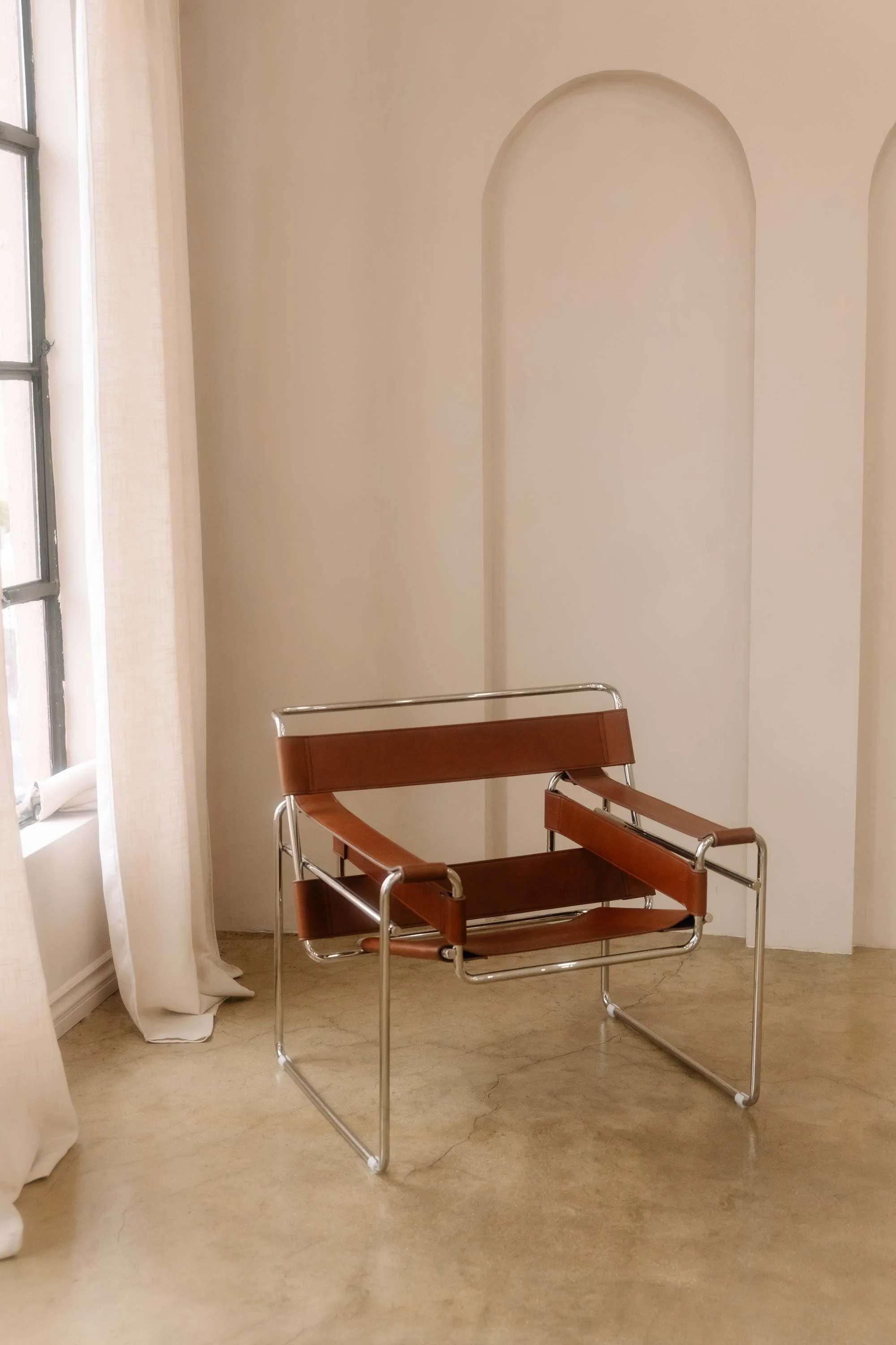 A modern chair with a brown leather and chrome frame is placed in a beige room with curved wall niches, near a window with white curtains.