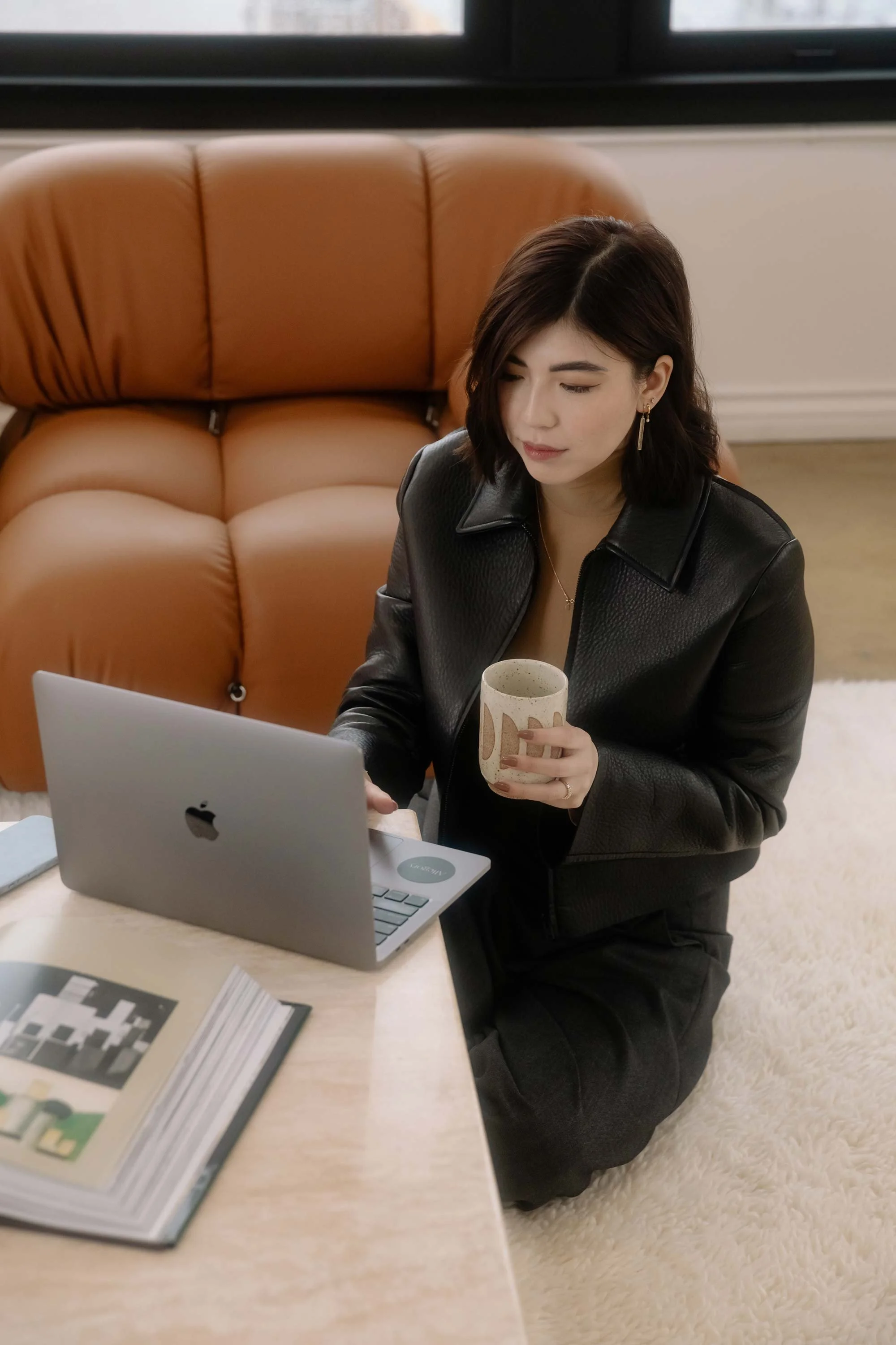 A woman kneeling on a white shag rug, working on a silver MacBook on a low table, holding a beige ceramic cup, with a brown sofa and a window in the background.
