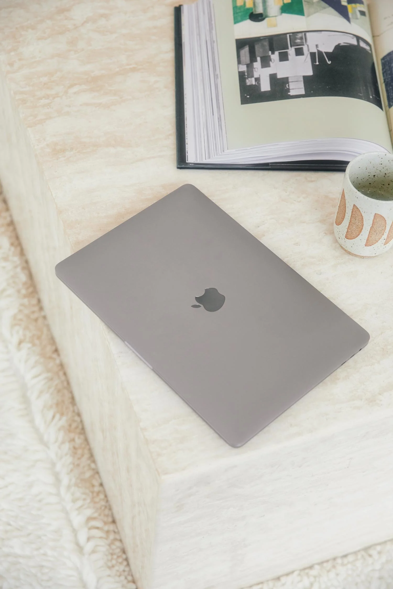 A closed silver MacBook laptop resting on a cream-colored surface, with an open book, a ceramic mug, and some papers nearby.