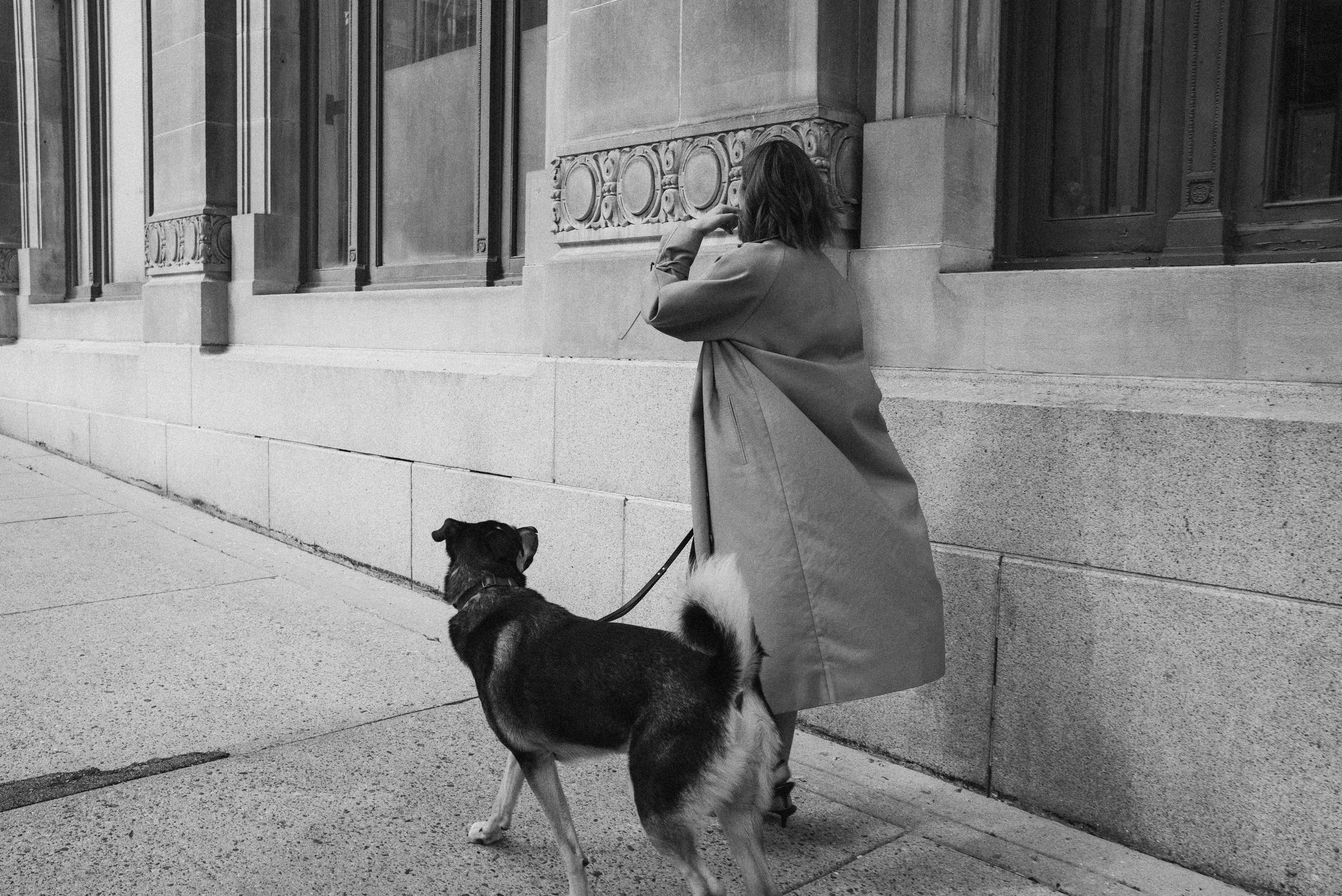 A woman in a long coat stands on a city sidewalk, talking on her phone, with a dog on a leash looking up at her, in front of a building with ornate windows.
