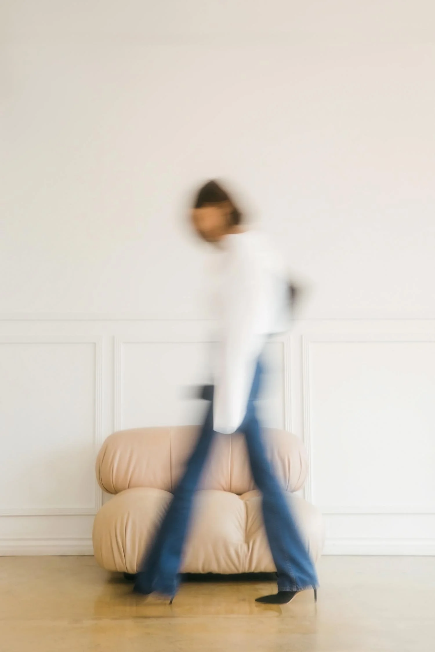 Blurred photo of a woman standing on a beige chair against a plain white wall, wearing a white top and blue jeans.