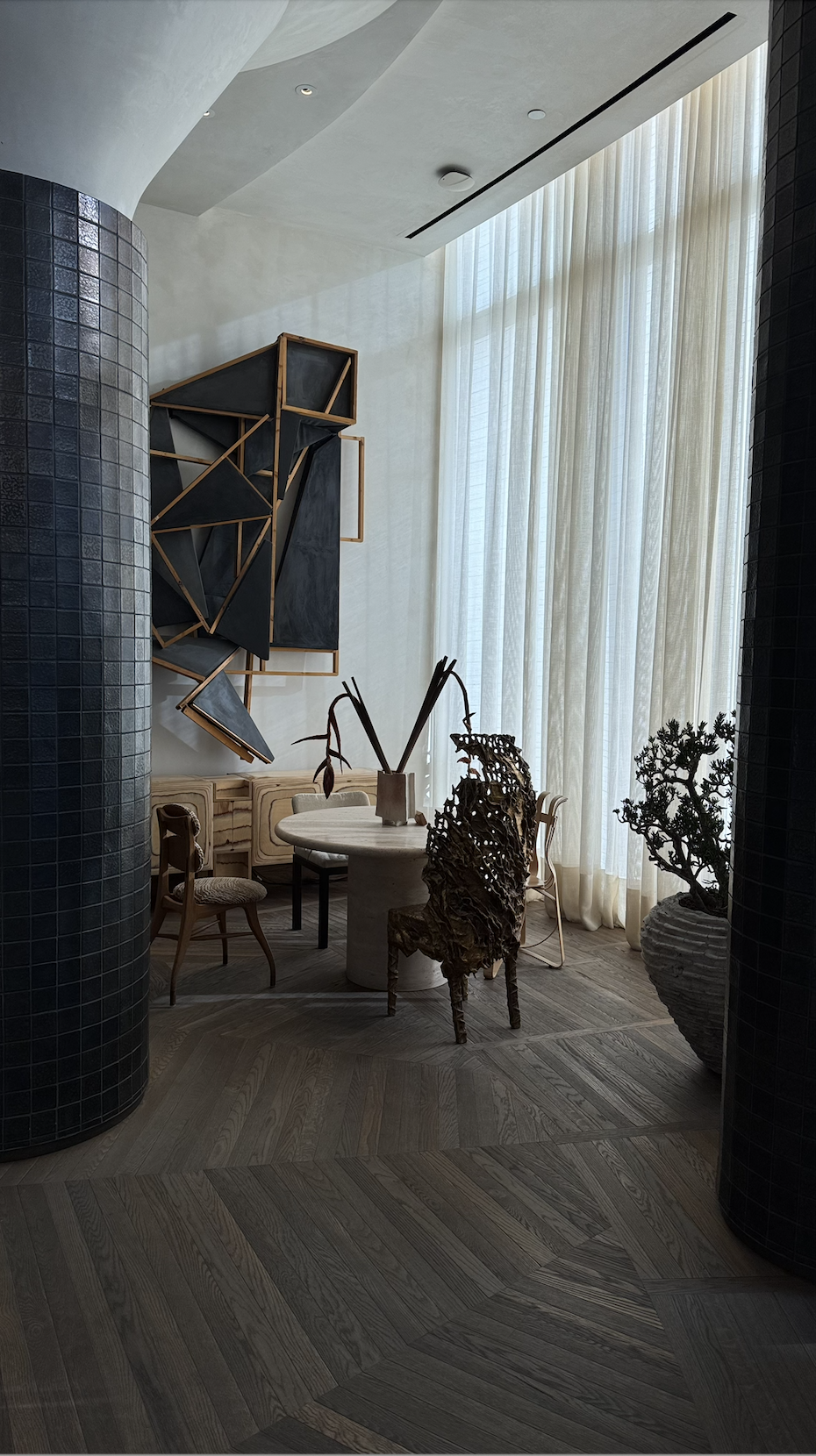 Interior of a modern room with a round table, various chairs, a large window with sheer curtains, and artistic wall decor.