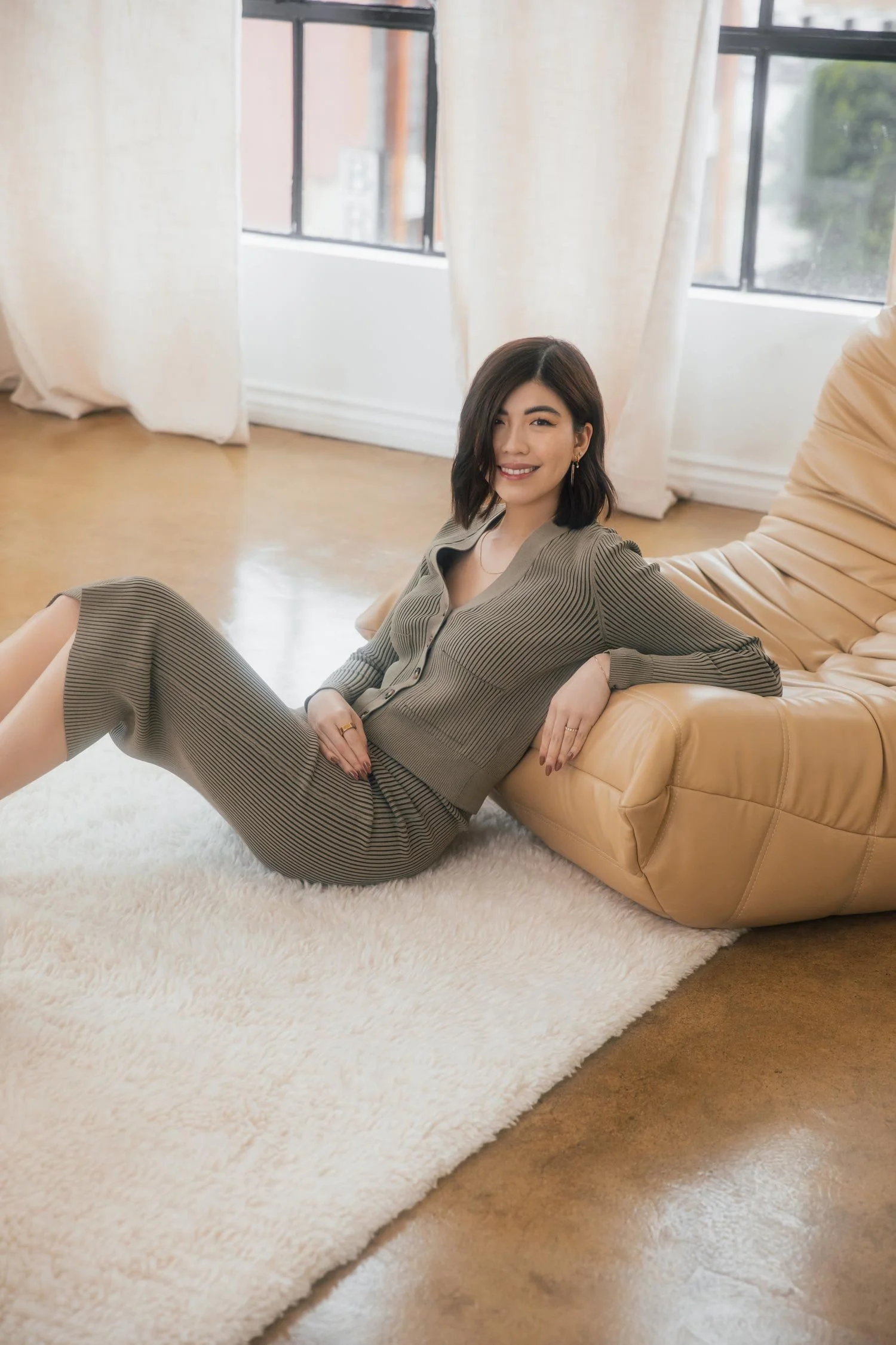 Tiffany Tang, founder of Allegory lounging on a beige leather chair, inside a bright room with large windows and white curtains.