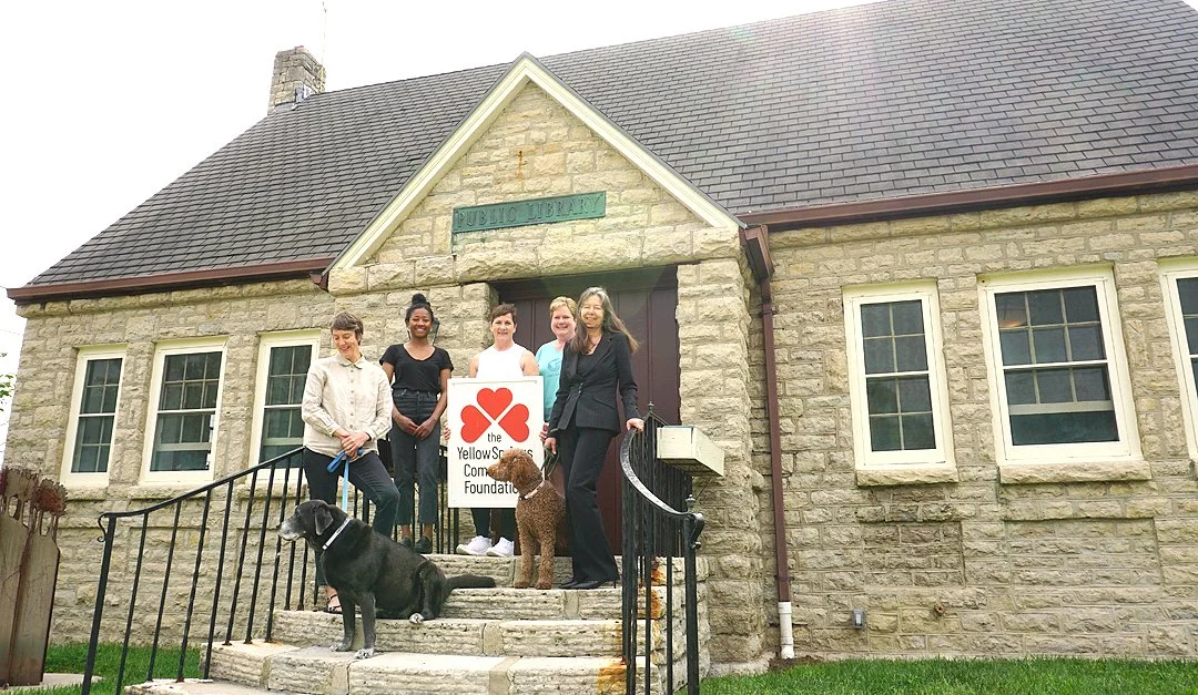 Group of five women standing on steps in front of a stone building labeled 'Public Library' holding a sign for the Yellow Springs Community Foundation, with a large black dog and a brown dog in front.