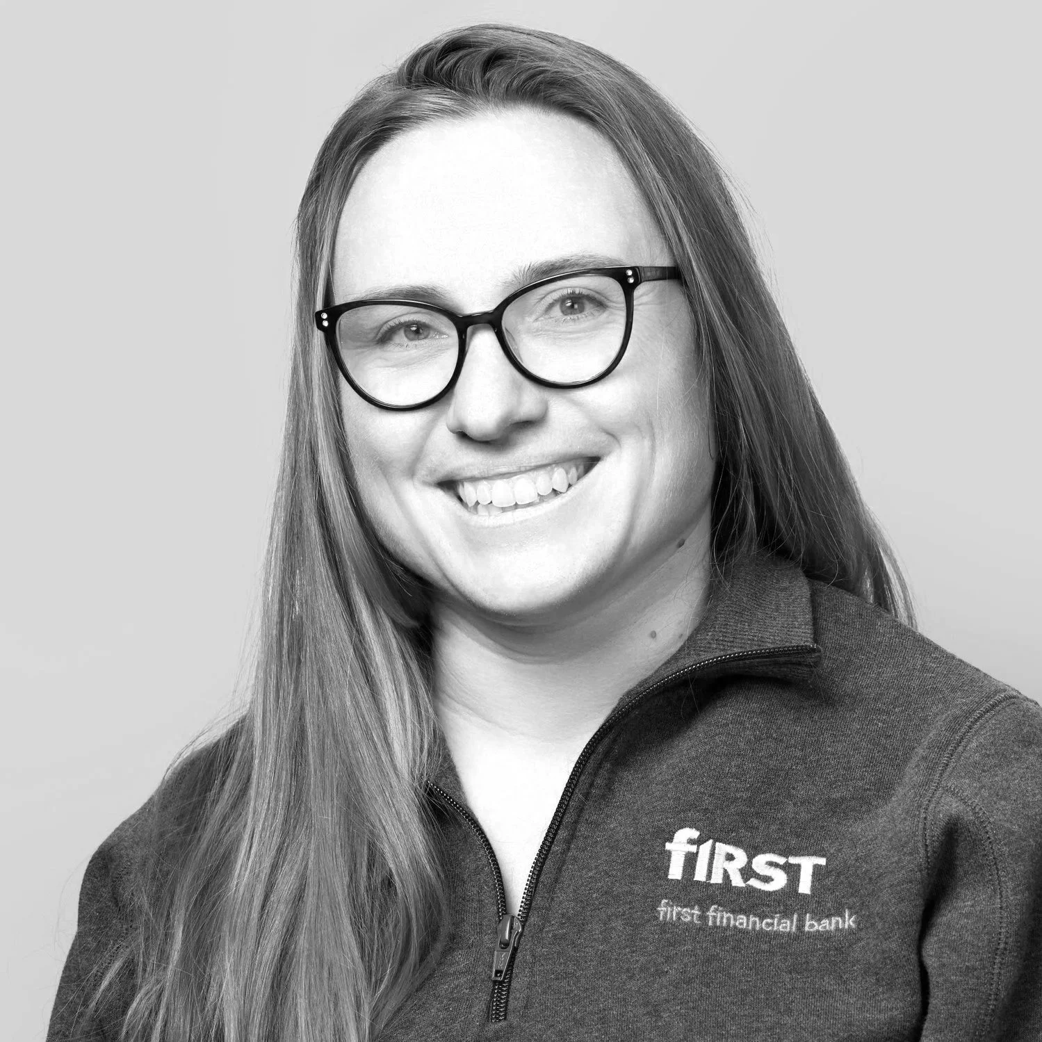 Black and white portrait of a woman with long hair, wearing glasses and a zip-up jacket with the First Financial Bank logo.