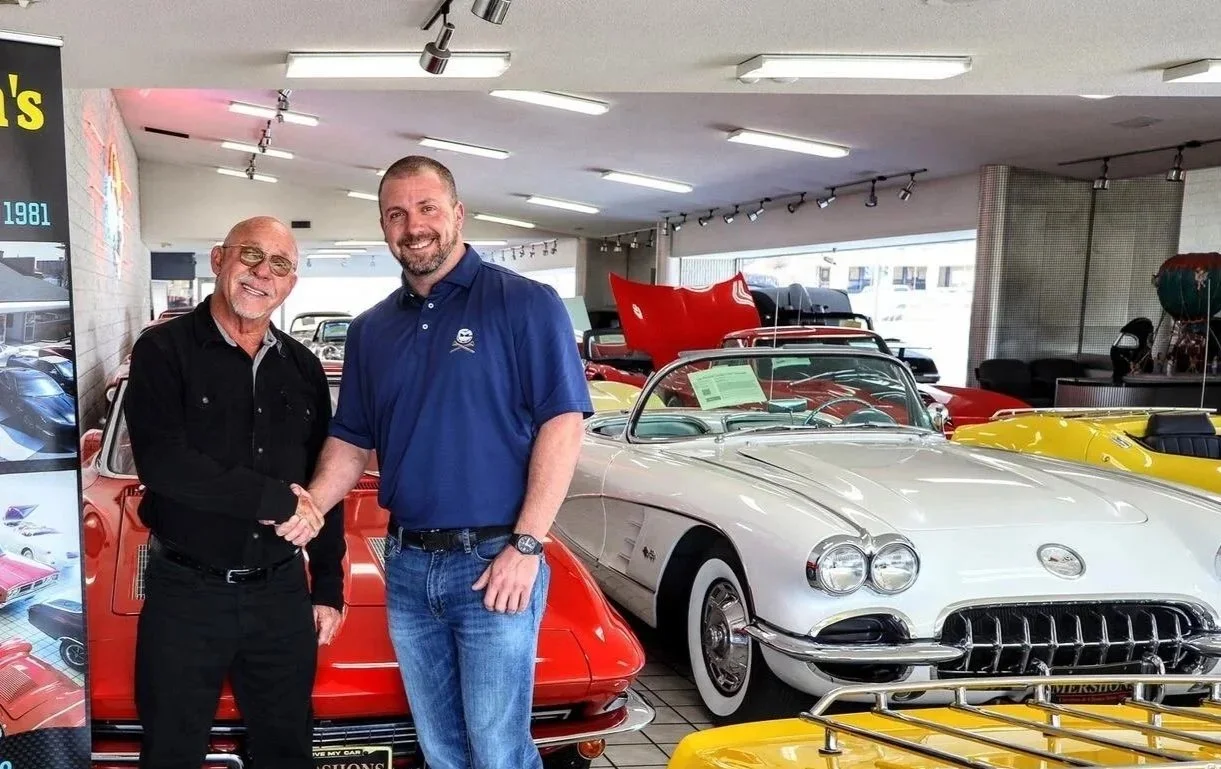 Two men shaking hands in a car museum filled with classic cars, including a red and a white convertible.