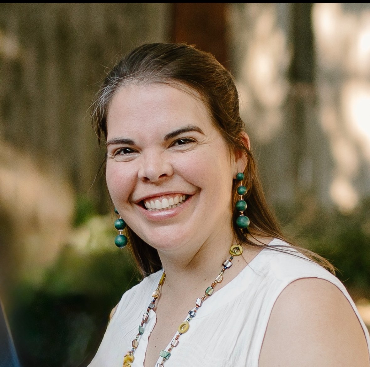 Elizabeth Wortham,, wearing a white sleeveless top, green earrings, and a colorful beaded necklace in an outdoor setting.