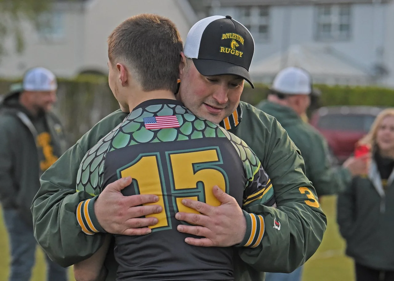 Two men hugging, one in a rugby uniform and the other in a jacket with the number 3, embracing after a game. The man in the rugby uniform wears a jersey with a large number 15, an American flag patch, and a dragon emblem. The background shows other people in jackets and caps, with a residential neighborhood behind them.