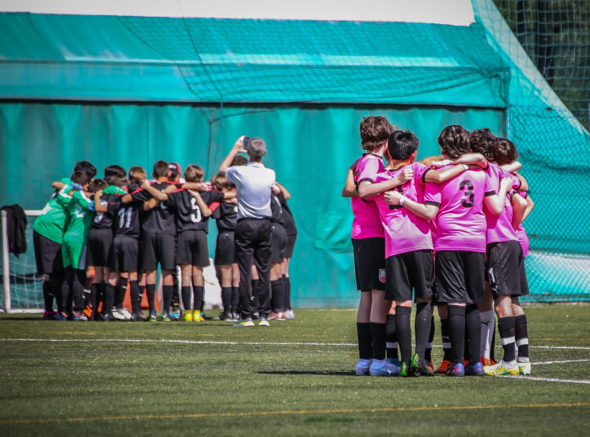 Two groups of young soccer players in pink and black uniforms huddled together on a soccer field during daytime