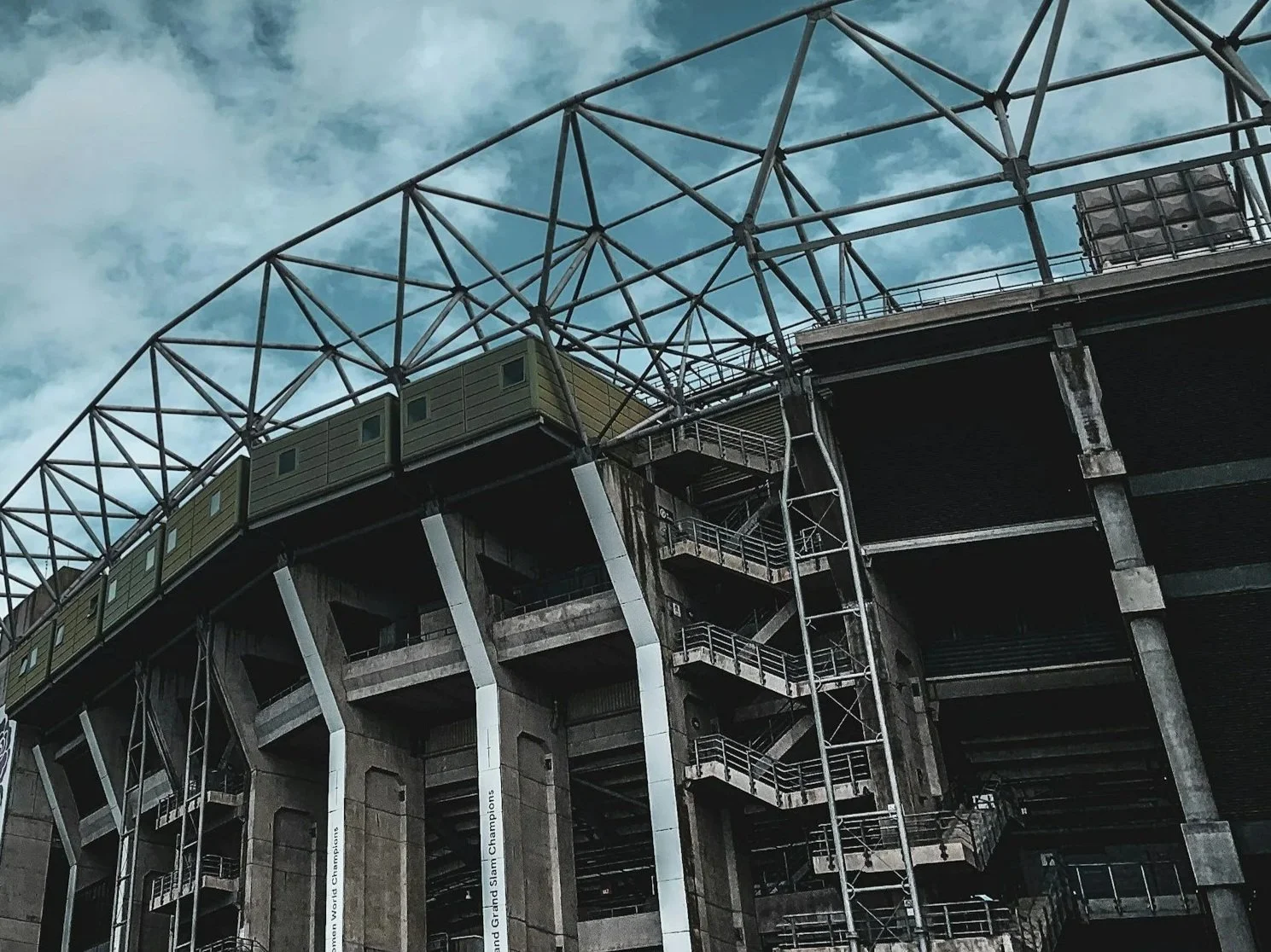 An industrial stadium structure with concrete columns, metal stairs, and a metal framework overhead under a cloudy sky.