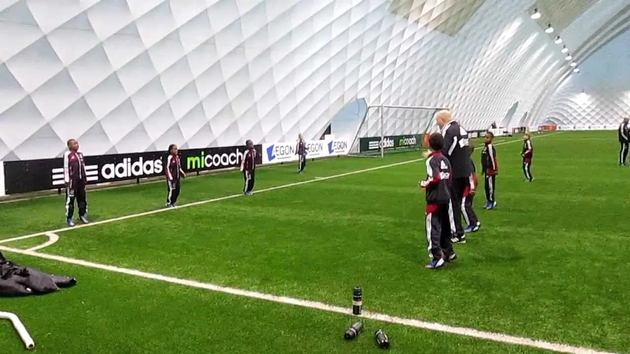 Children and an adult practicing soccer inside a modern indoor sports facility with a curved, textured ceiling. The children are lined up near the goal, and the adult appears to be instructing them.