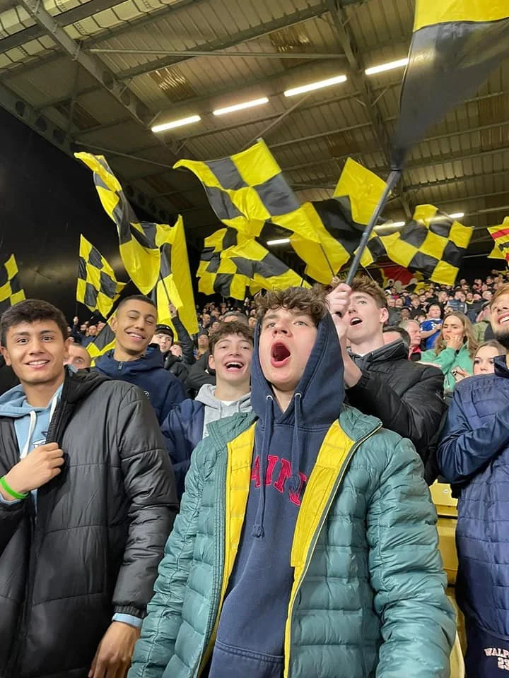 Group of young people at a stadium holding yellow and black checkered flags and cheering for a team.