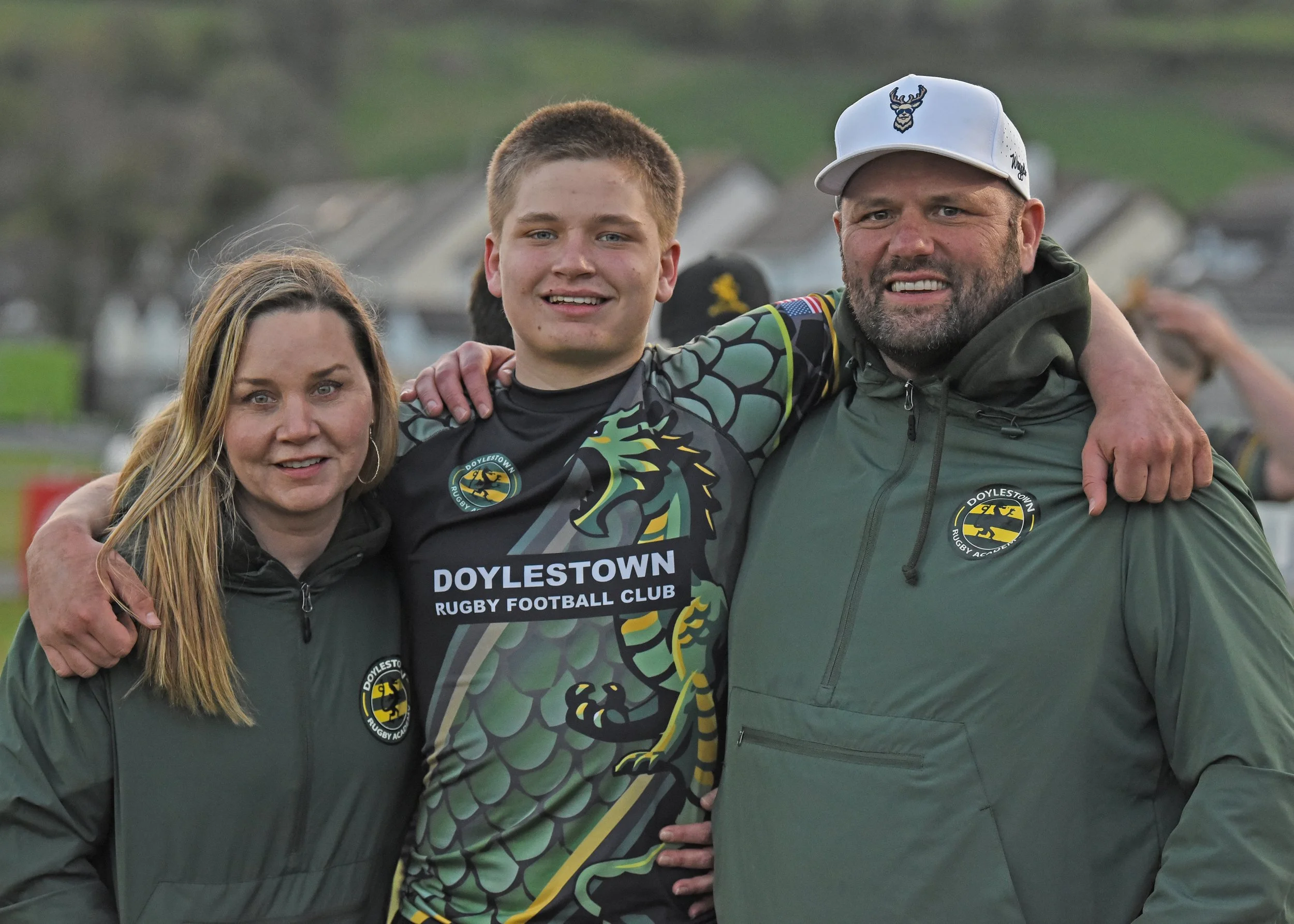 Three people standing outdoors, smiling, with a house and trees in the background. They are wearing Doylestown Rugby Football Club gear, with the young man in the center wearing a black and green rugby jersey and the two adults in green jackets. The people are posing closely together with their arms around each other.