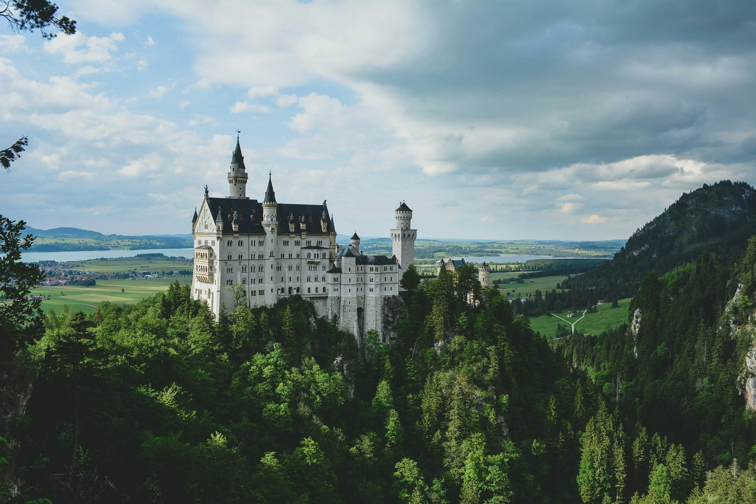 A large white castle with tall towers and black rooftops, situated on a hill surrounded by dense green trees, with a cloudy sky overhead and a vast landscape in the background.