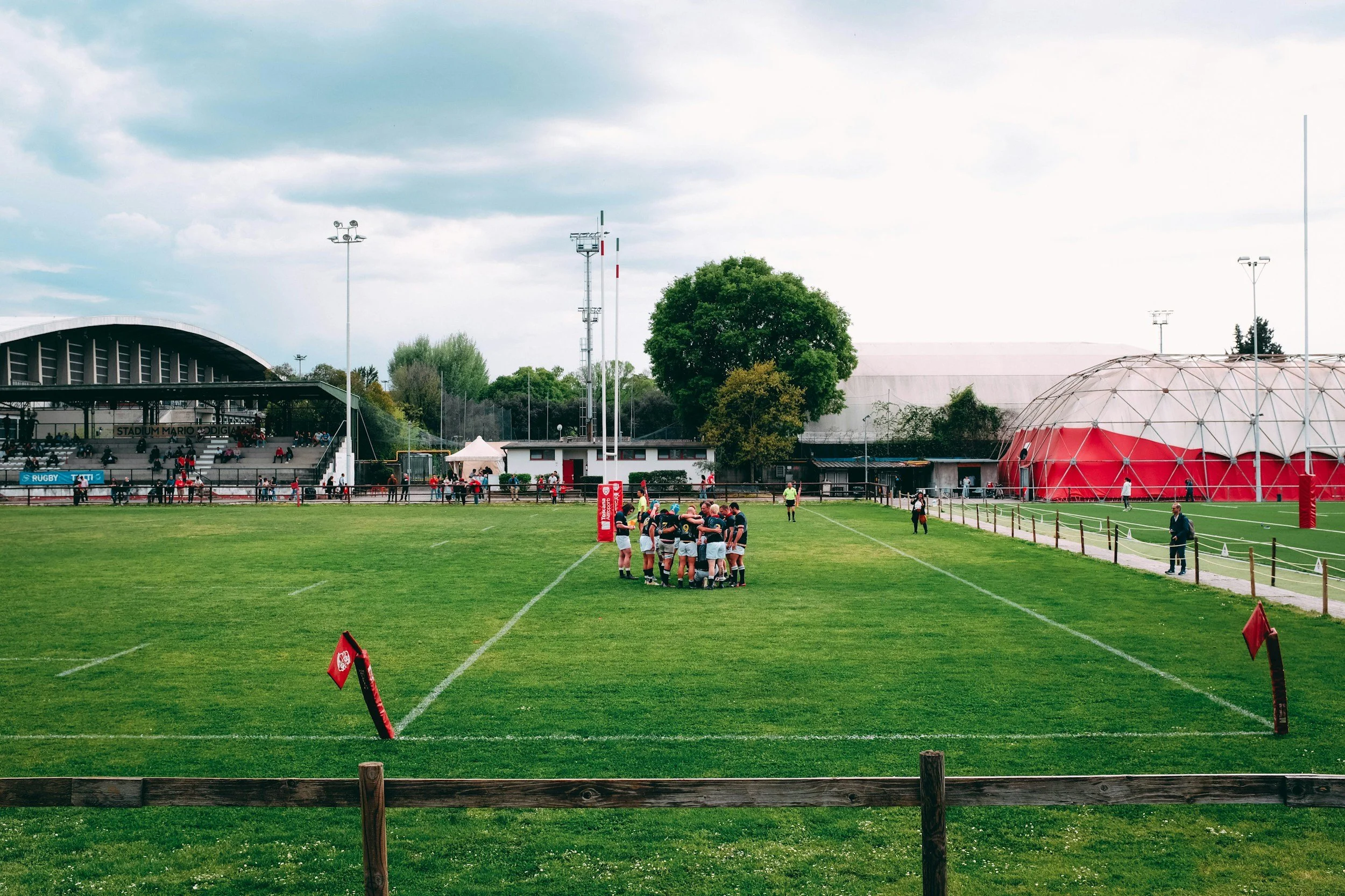 Rugby players huddled together on a grassy field during a match at a sports stadium with spectators in the stands and a large dome-shaped structure in the background.
