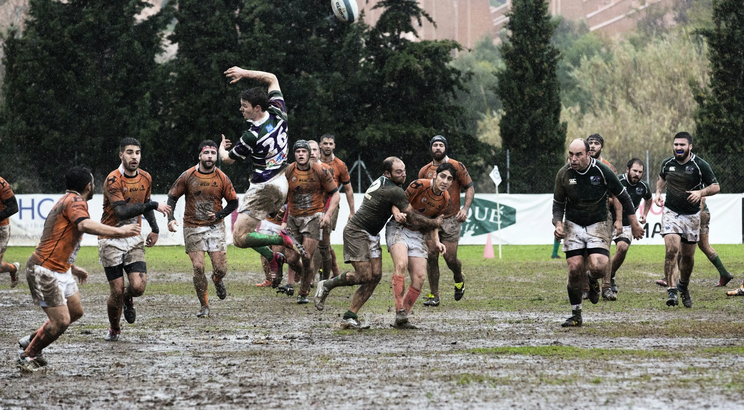 Rugby players on a muddy field during a game, with one player jumping to catch the ball.