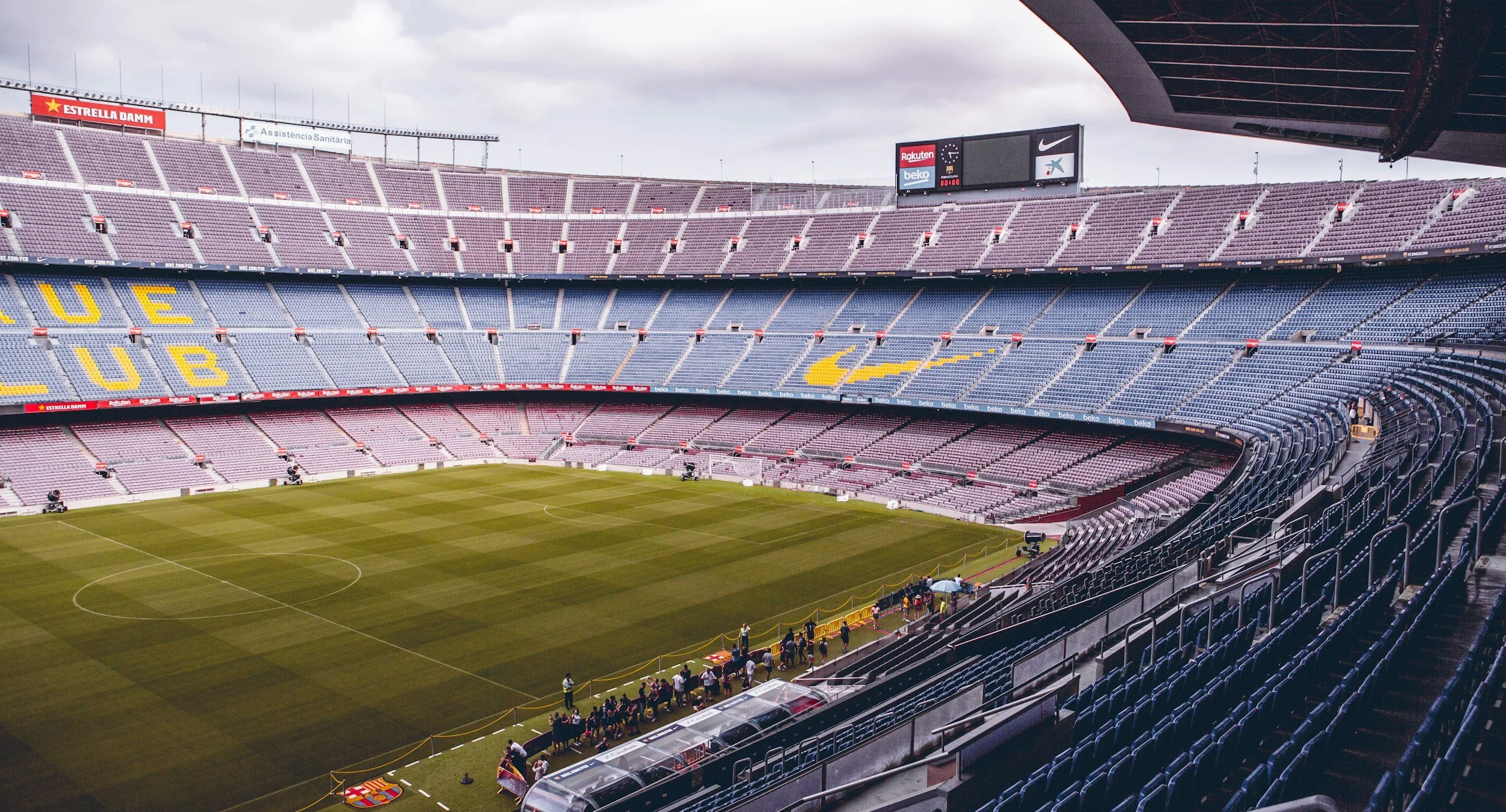Empty soccer stadium with a green field, surrounded by multiple tiers of seating stands, some with seats and some empty. Overcast sky above, scoreboard in the background, and a few people near the sideline.