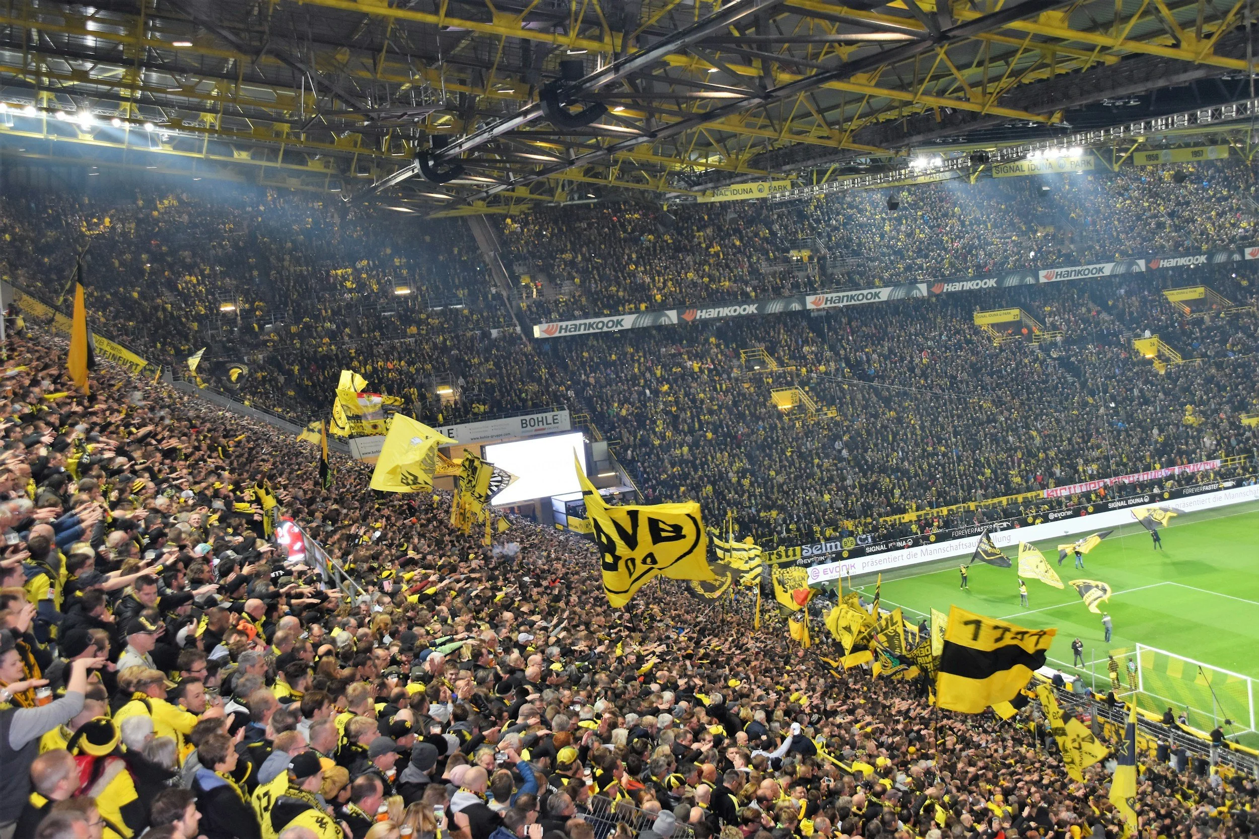Crowd of football fans in yellow and black jerseys inside a stadium, waving flags and cheering during a match.