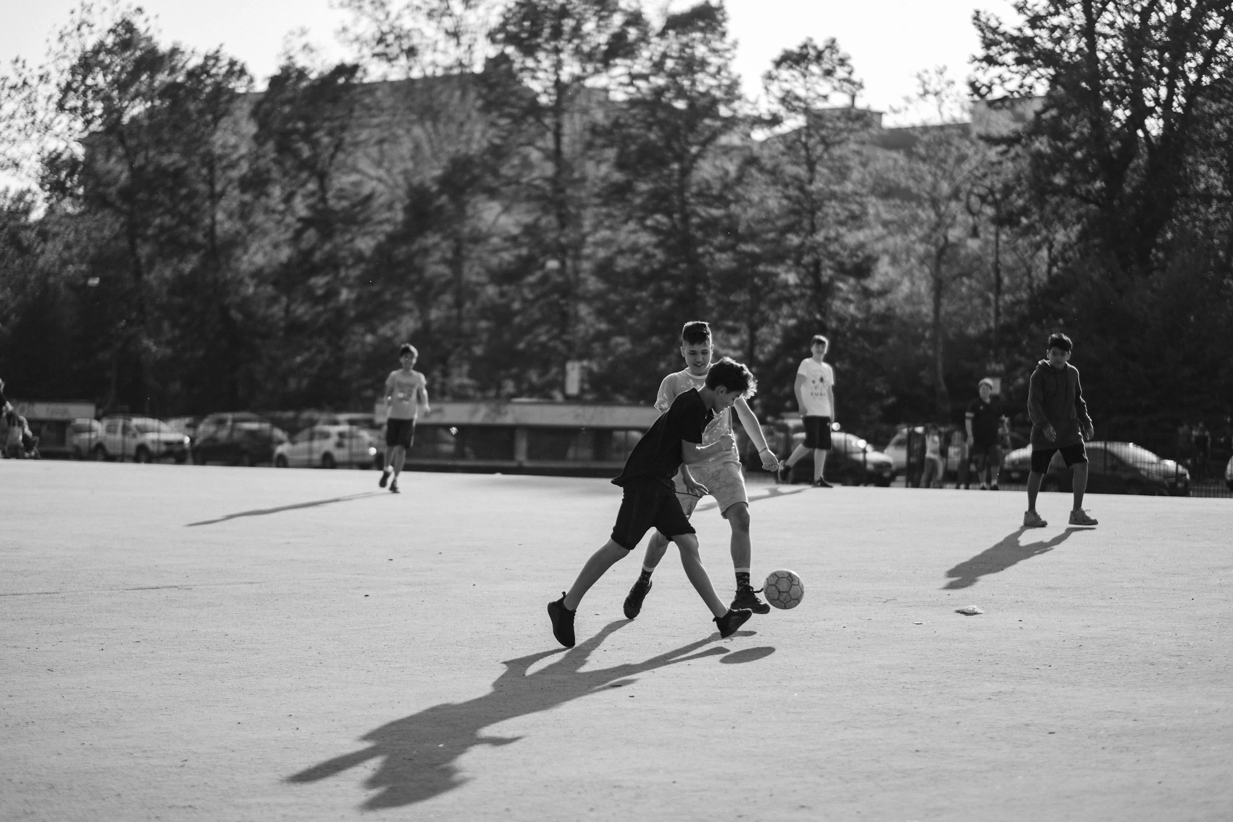 Young boys playing soccer on an outdoor field with trees and parked cars in the background, depicted in black and white.