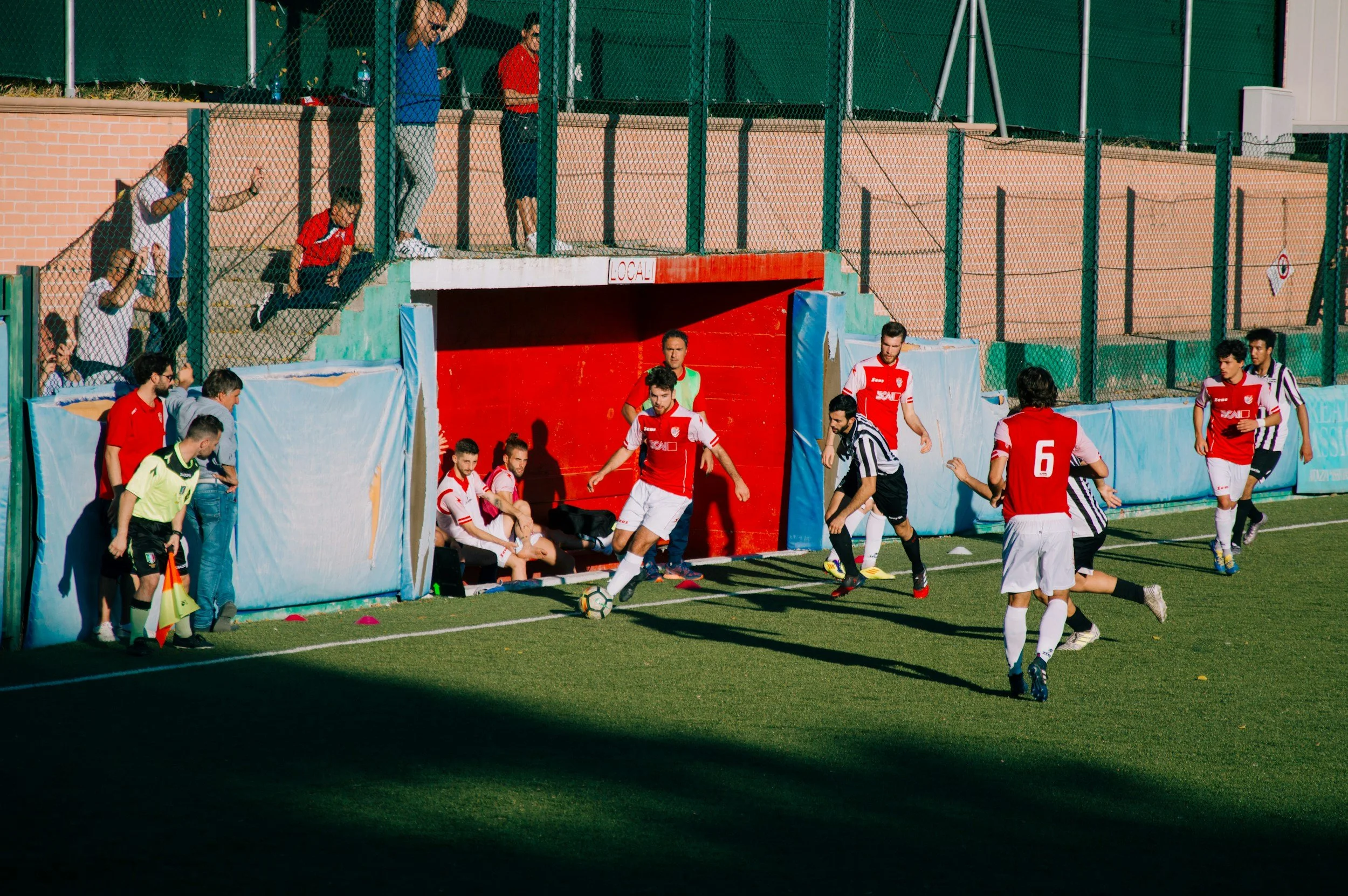 Soccer match with players near the bench on field, some players sitting on bench, others running or standing, some spectators watching from stands behind a fence in sunny weather.