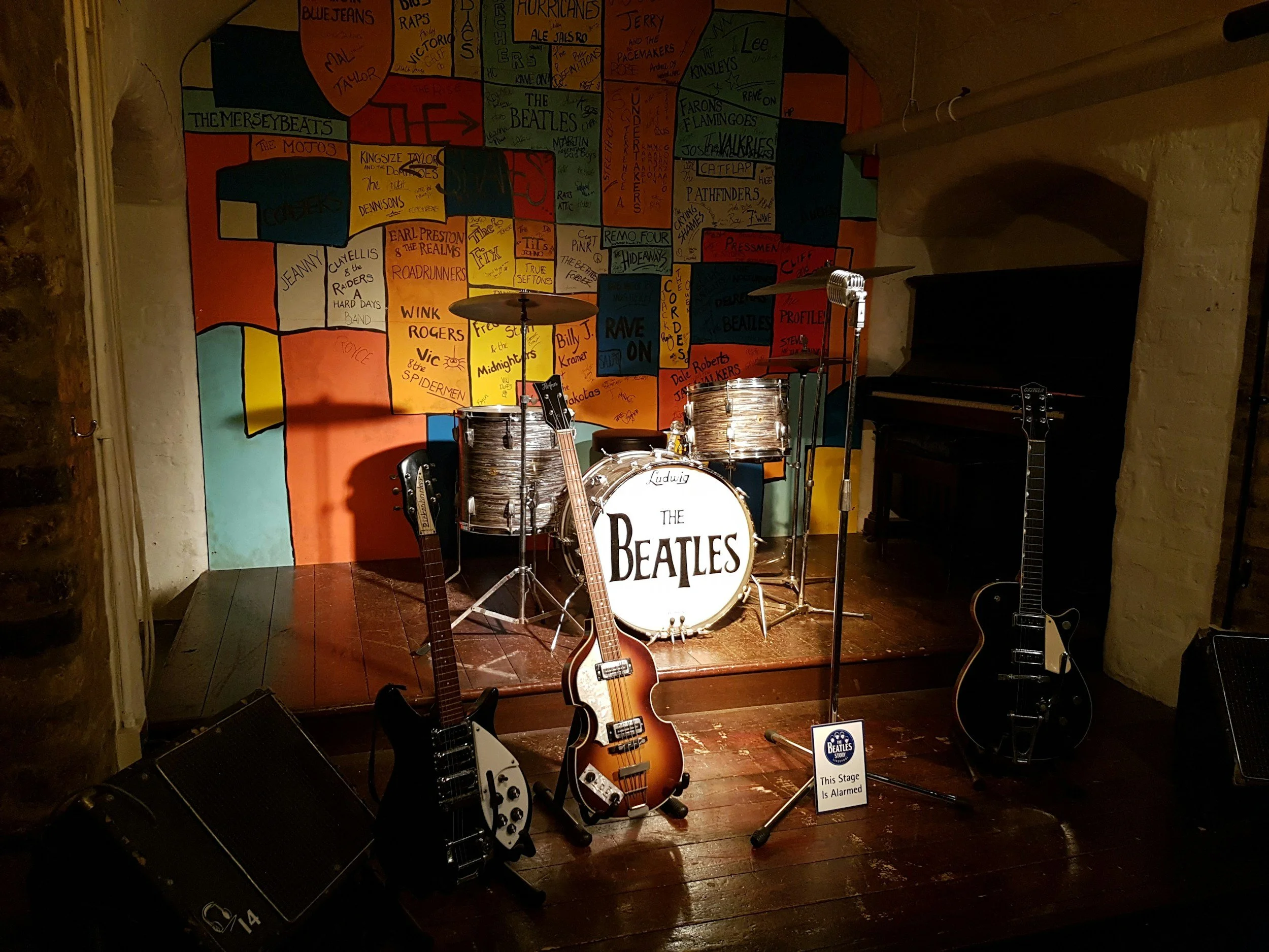A stage decorated with a Beatles theme, featuring a drum set, several electric guitars, a sign that says "This Stage is Alarmed," and a colorful mural of handwritten band names and song titles on the wall behind.
