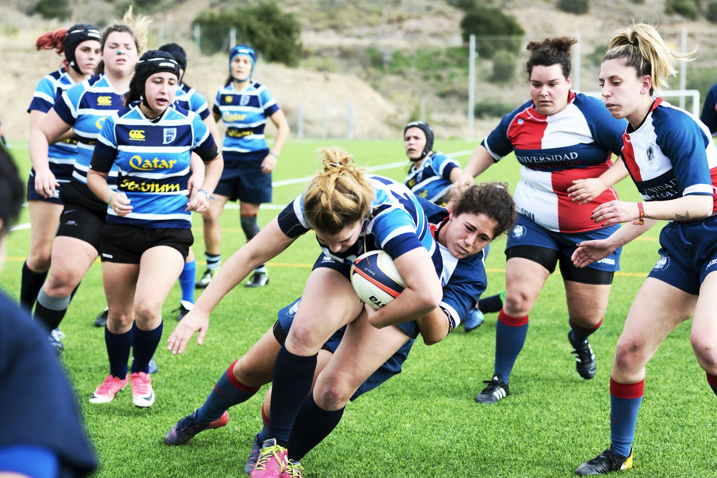Women playing rugby on a grassy field, with some players wearing blue and striped jerseys and others wearing red, white, and blue jerseys. One woman is running with the ball while others are trying to block her or support.
