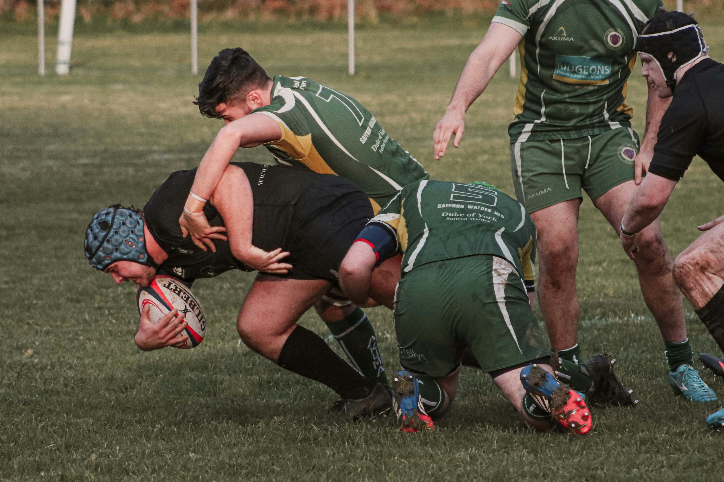 Rugby players engaged in a tackle on the field, with one player holding the rugby ball and others attempting to stop him.